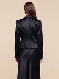 A woman with long brown hair, seen from behind, wears the Misa Blazer in black pinstripe with a matching satin suiting skirt, standing against a plain, light background.