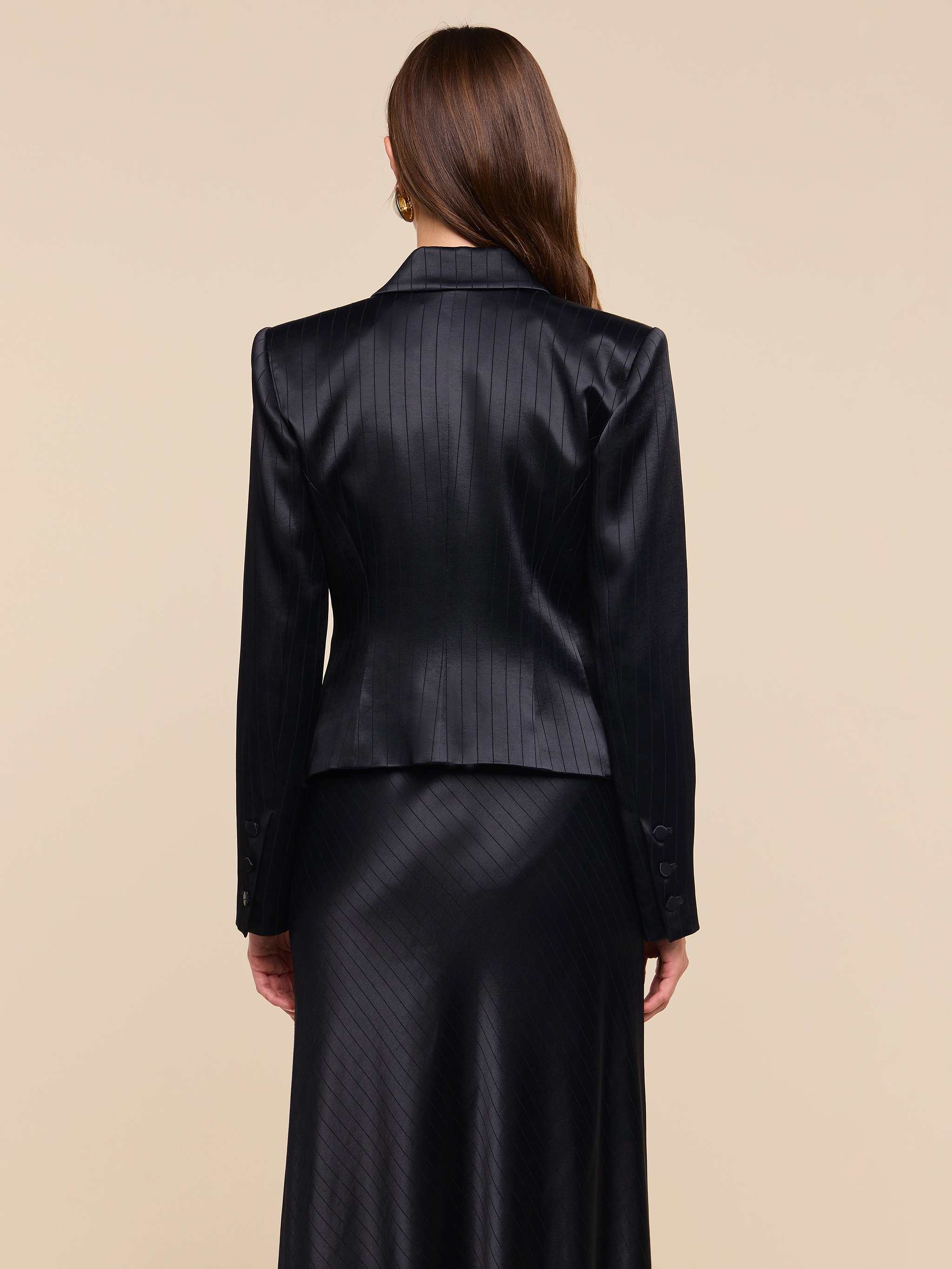 A woman with long brown hair, seen from behind, wears the Misa Blazer in black pinstripe with a matching satin suiting skirt, standing against a plain, light background.