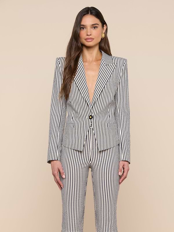 A woman stands against a plain background, wearing the Misa Seersucker Blazer—a black and white vertical striped blazer with a single button—open at the chest, her long hair down, and accessorized with gold earrings.