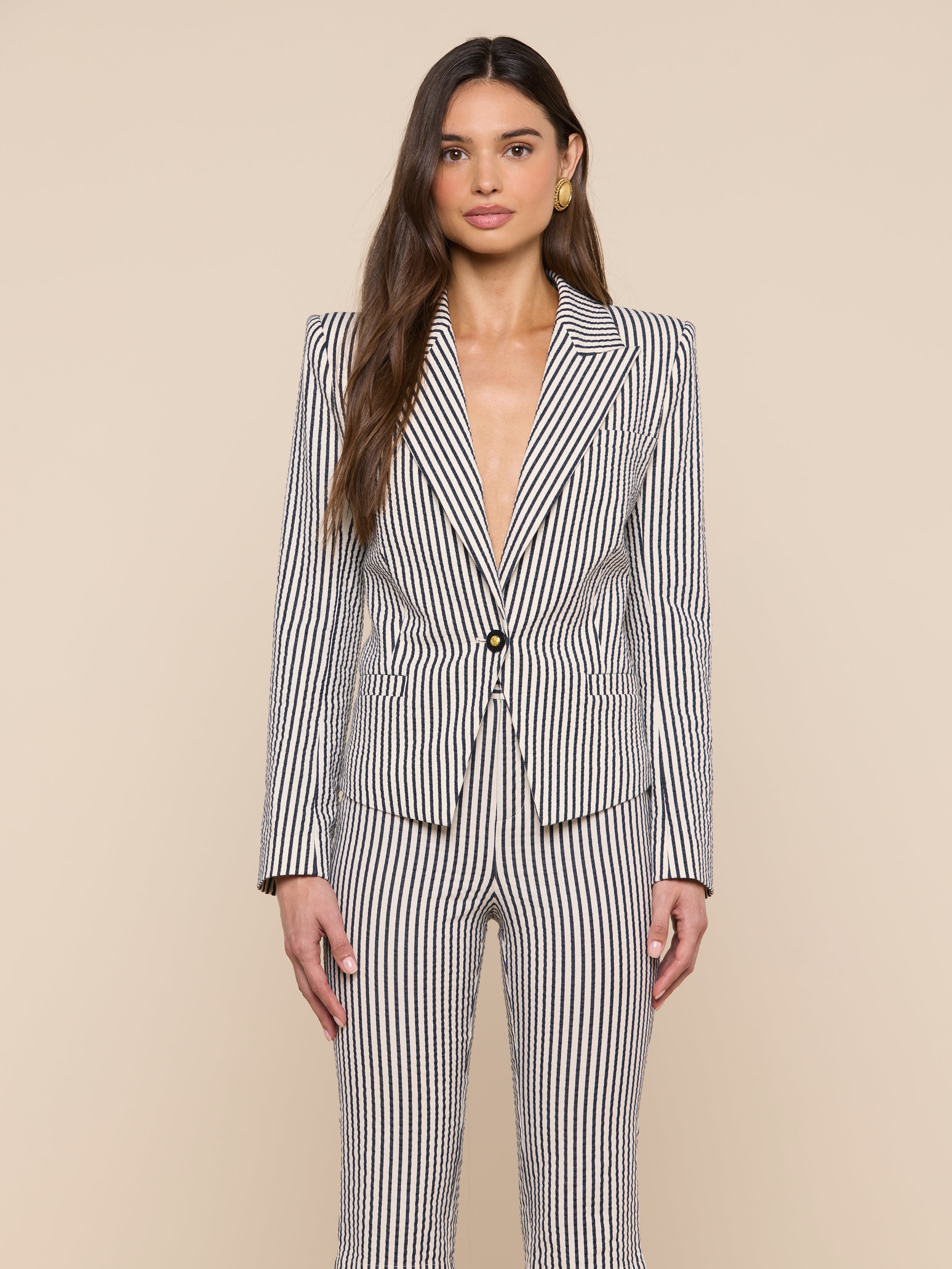 A woman stands against a plain background, wearing the Misa Seersucker Blazer—a black and white vertical striped blazer with a single button—open at the chest, her long hair down, and accessorized with gold earrings.