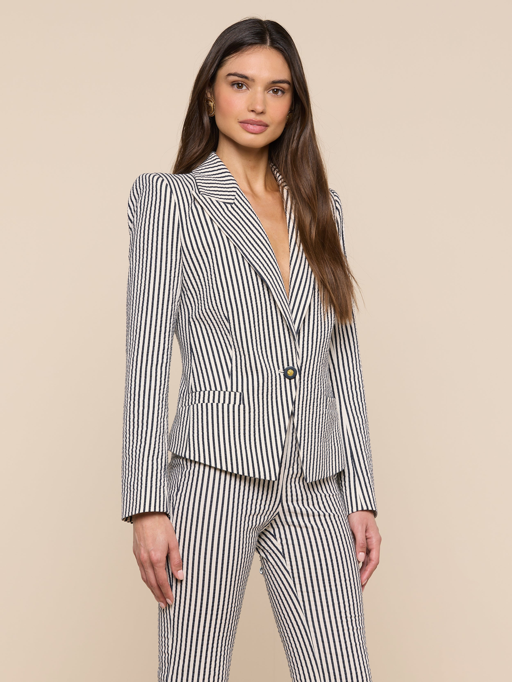 A woman stands facing forward against a plain light background, wearing the Misa Seersucker Blazer—a white blazer featuring black vertical stripes. She has long brown hair down and a neutral expression.