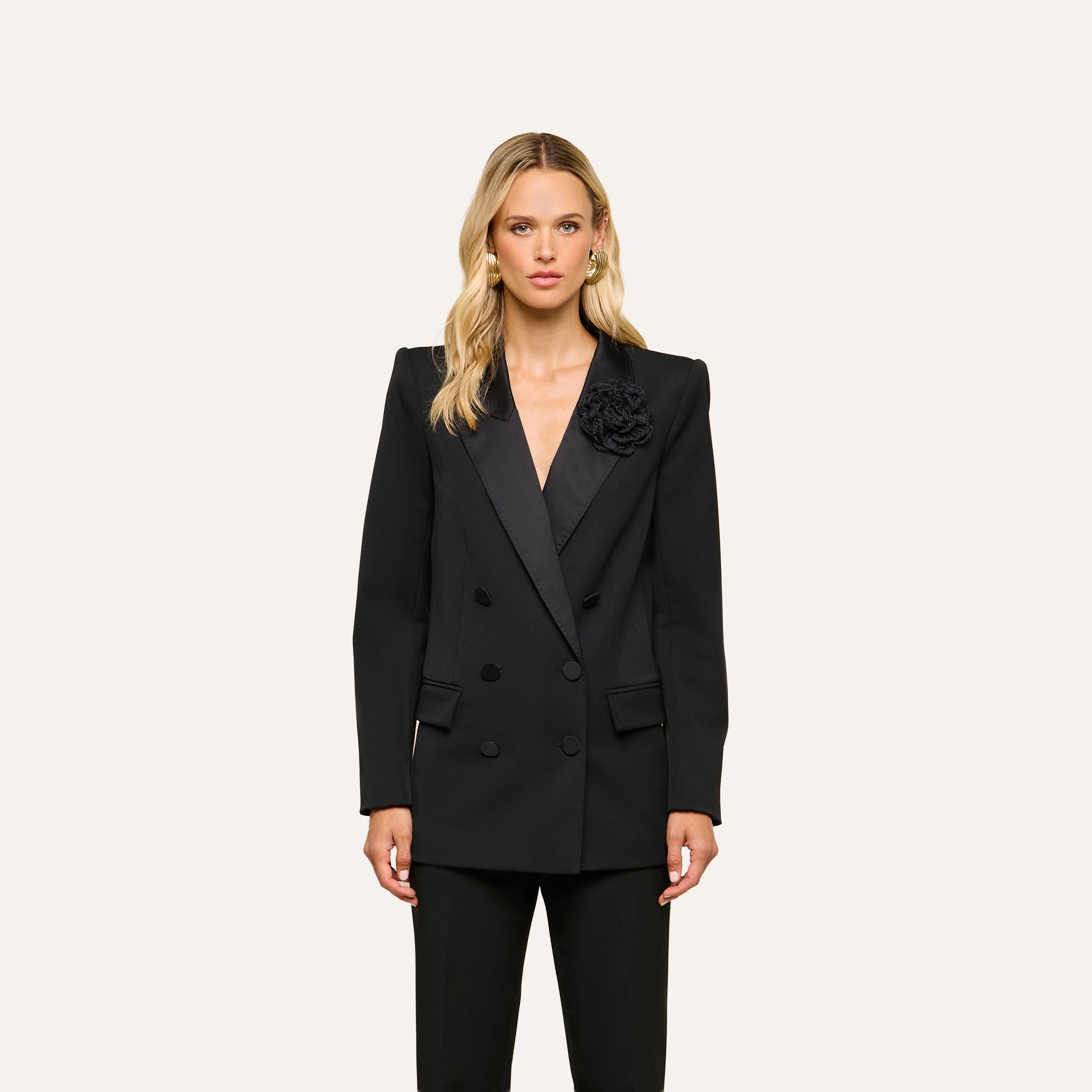 A person with long blonde hair wears the Cambrie Oversized Blazer, a black double-breasted boyfriend blazer featuring a flower detail, standing against a plain light background.