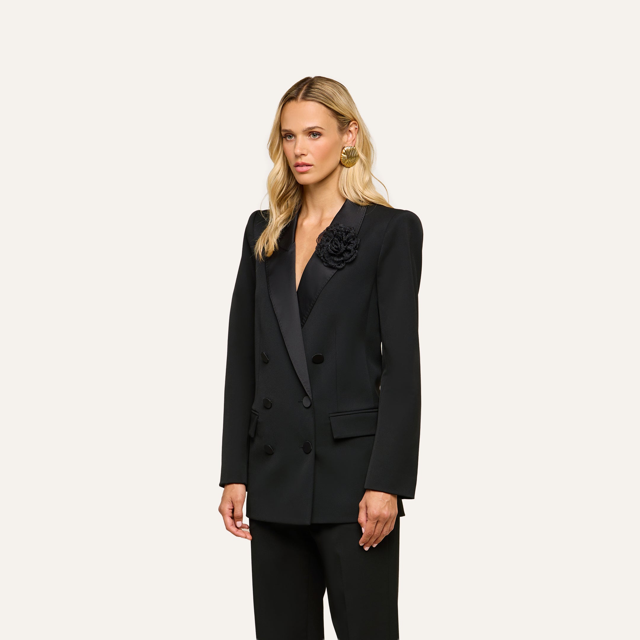 A woman with long blonde hair wears the Cambrie Oversized Blazer, featuring a double-breasted design and a flower detail on the lapel, paired with matching black trousers as she stands against a plain light background.