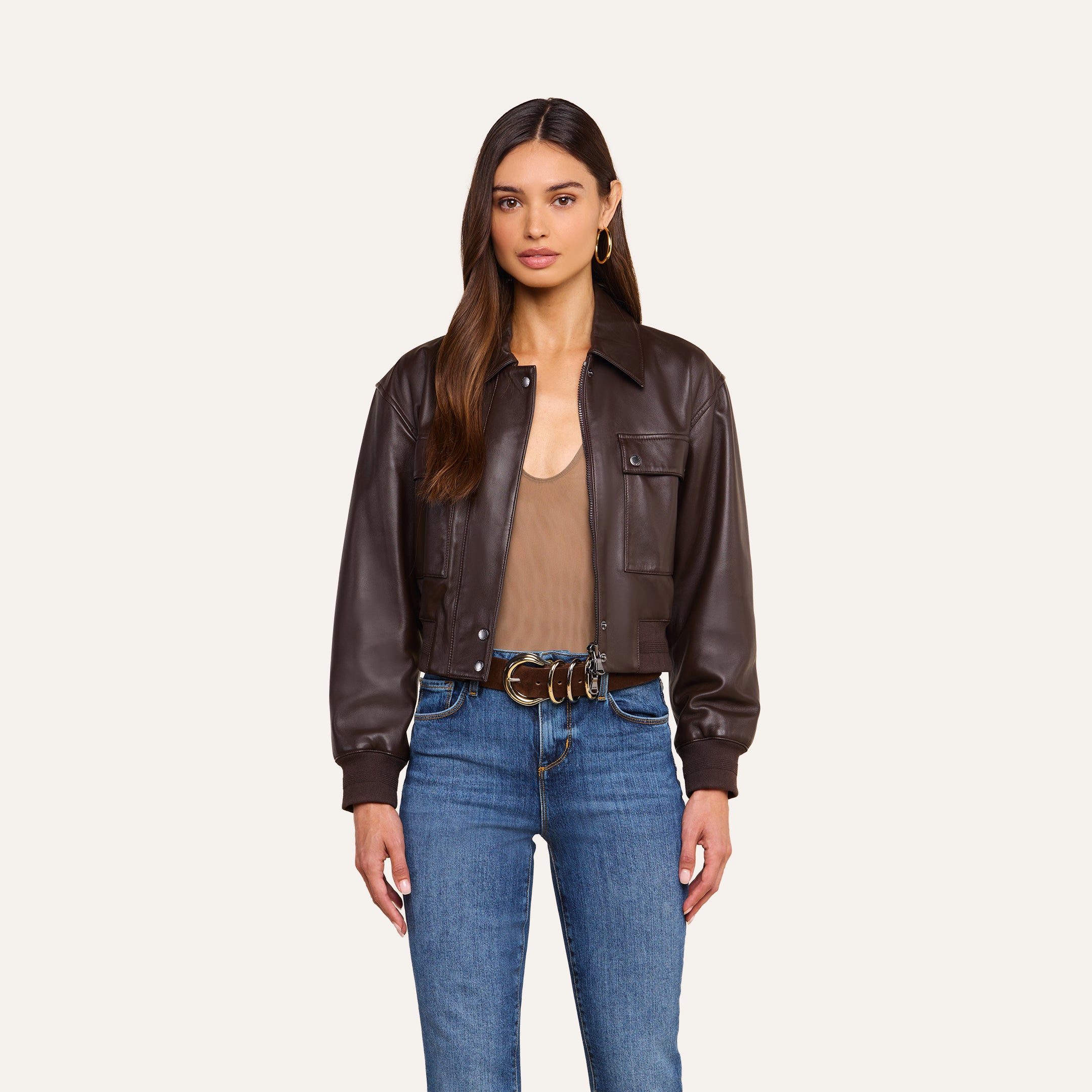 A woman with long brown hair wears the Clotilde Leather Jacket, styled with a tan top, blue jeans, and a belt. She stands facing forward against a plain light background.