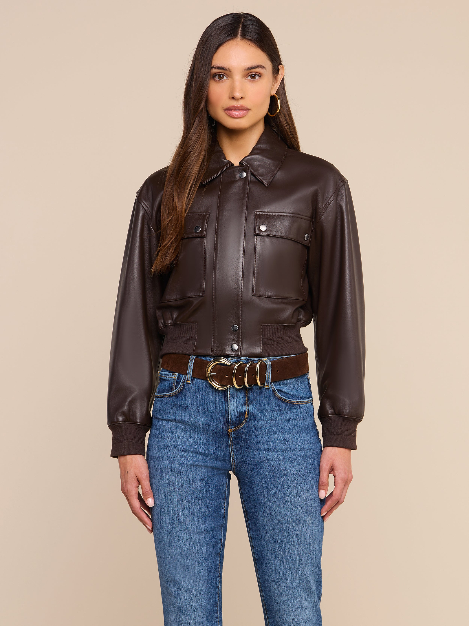 A woman stands facing forward, wearing the Clotilde Leather Jacket—crafted from fine-grain leather—paired with blue jeans and a brown belt with a gold buckle, set against a plain light background.