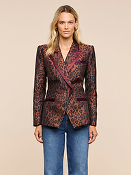 A woman with blonde hair and a neutral expression stands against a plain background, wearing the Kyomi Double-Breasted Blazer — a brown and burgundy leopard jacquard style with dark lapels — paired with blue jeans.