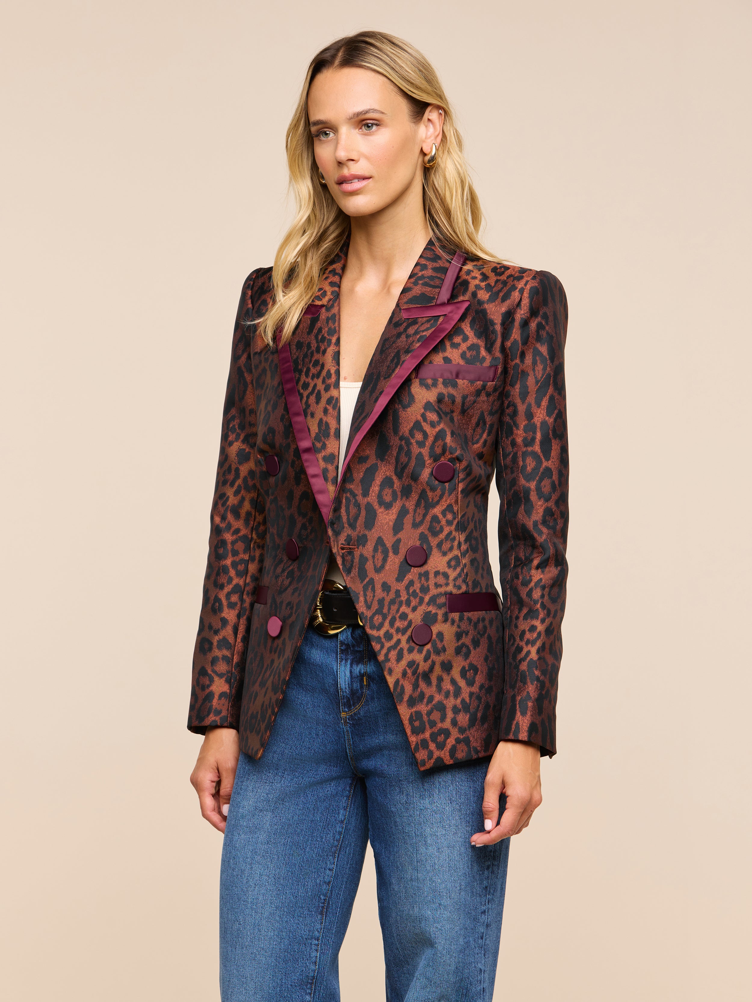 Wearing the Kyomi Double-Breasted Blazer with leopard jacquard and maroon trim over a light top and blue jeans, a woman stands against a plain light background.