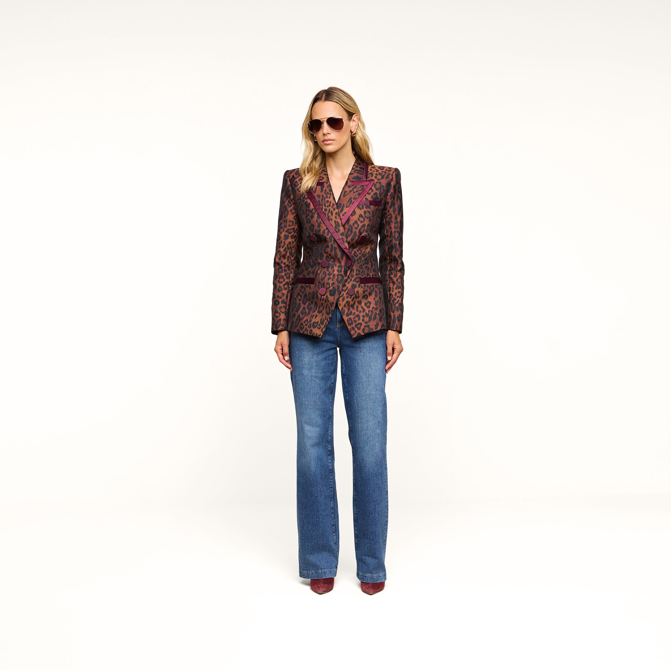 A person wearing sunglasses, the Kyomi Double-Breasted Blazer in leopard jacquard, blue jeans, and brown shoes stands against a plain white background.