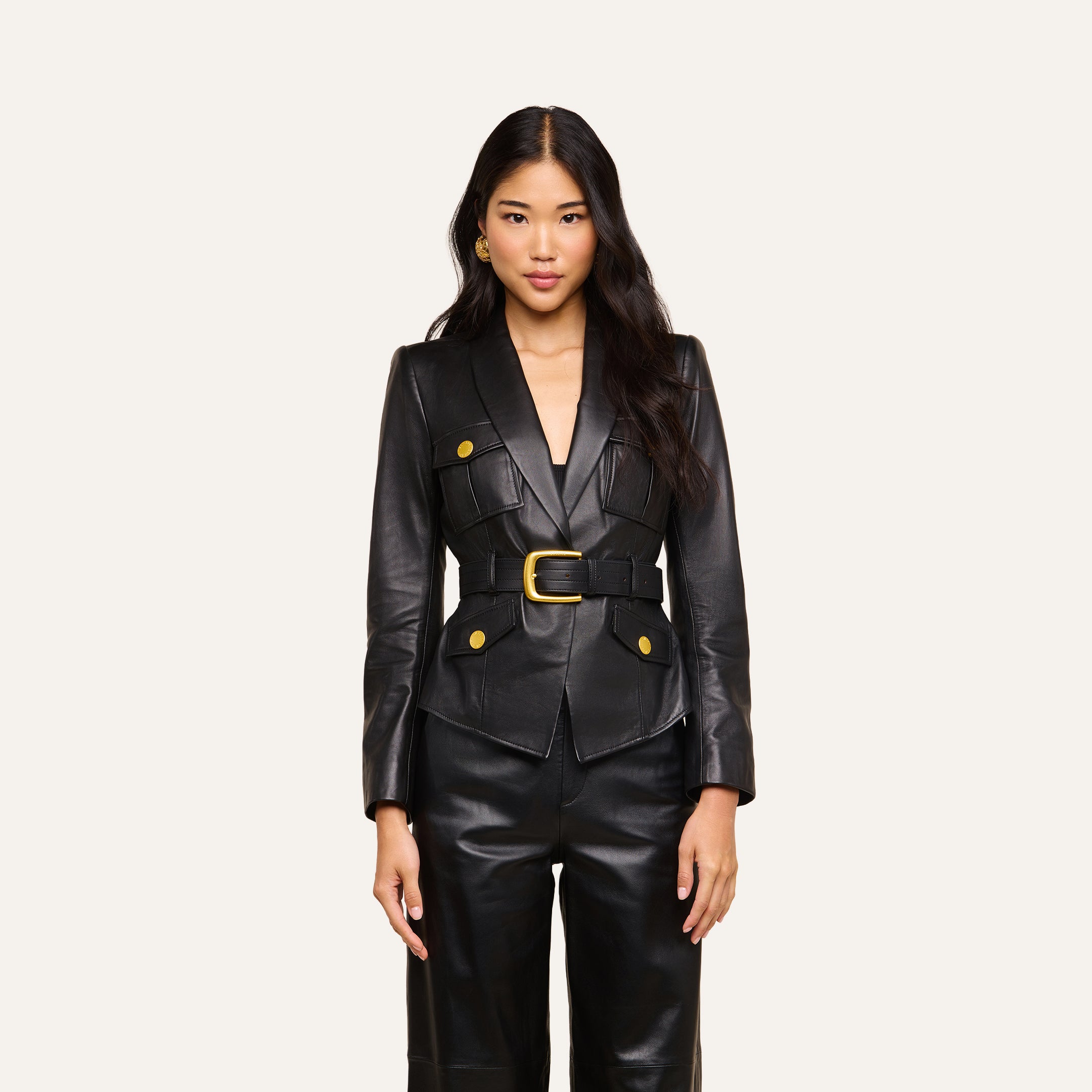 A woman stands facing forward against a plain light background, wearing the Wisteria Leather Blazer—a fitted black leather blazer with gold buttons and a belt—paired with matching black leather pants.