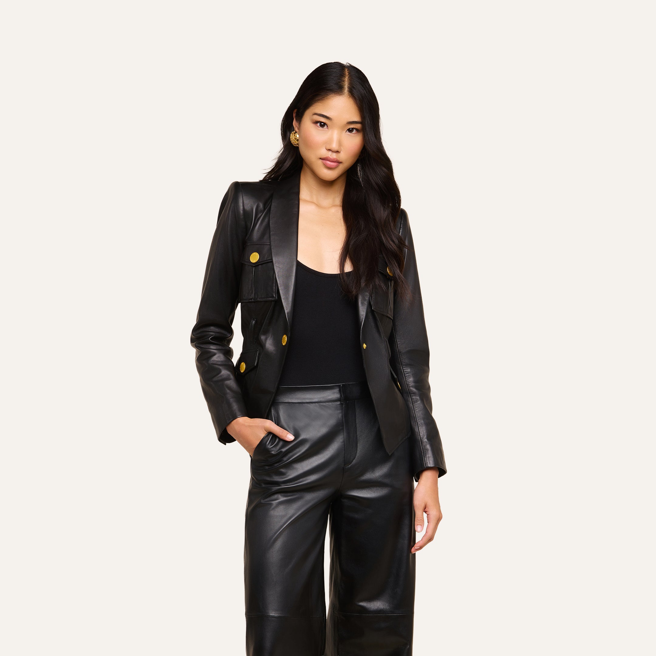 A woman with long black hair wears the fitted Wisteria Leather Blazer with gold buttons, paired with a black top and matching black leather pants, standing with one hand in her pocket against a plain light background.