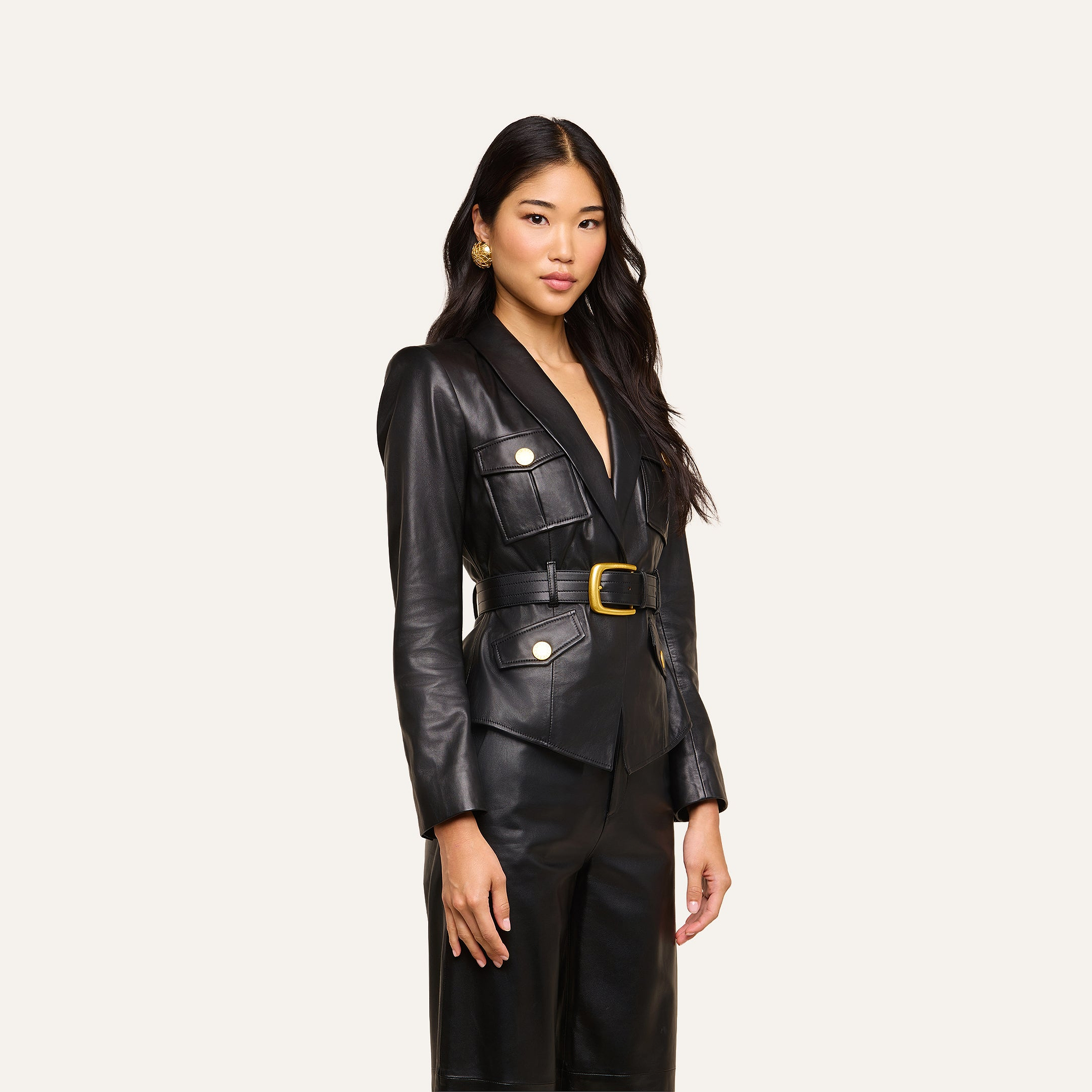 A woman with long dark hair wears the Wisteria Leather Blazer—a fitted black leather jacket with gold buttons—paired with matching pants, standing confidently against a plain light background.