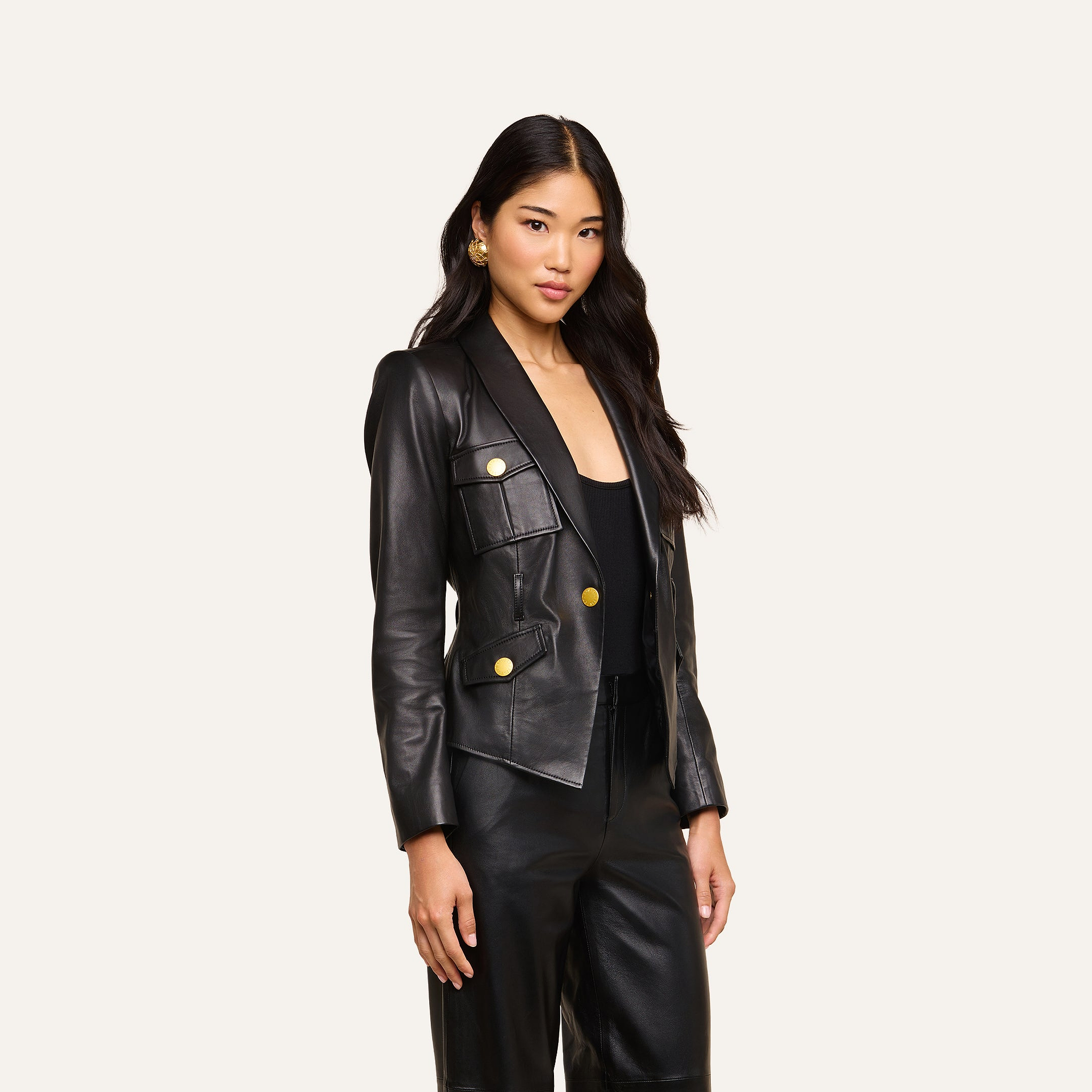 A woman with long, wavy hair wears the fitted Wisteria Leather Blazer with gold buttons and matching black leather pants, standing against a plain light background.