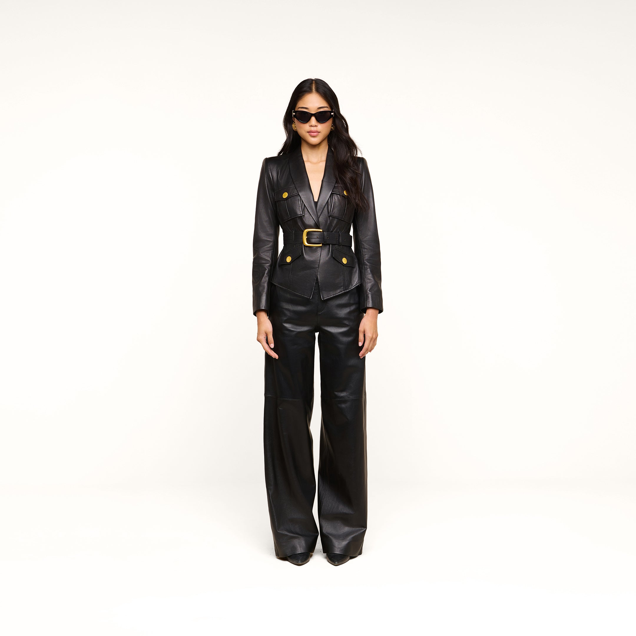 A woman wearing the Wisteria Leather Blazer with gold buttons, wide-leg black leather pants, and sunglasses stands against a plain white background.