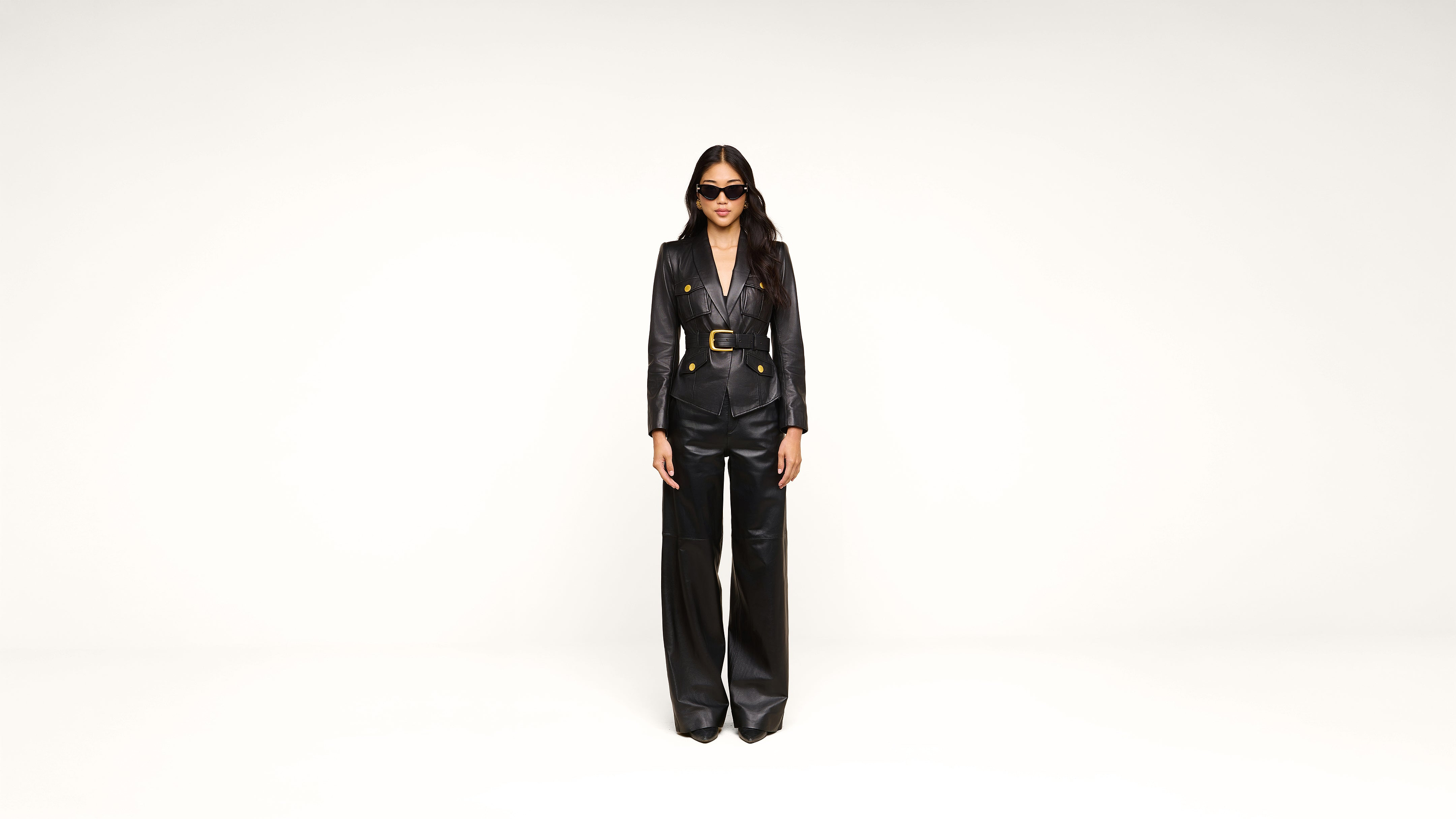 A person wearing black sunglasses, a black belted blazer, and black wide-leg pants stands against a plain white background.