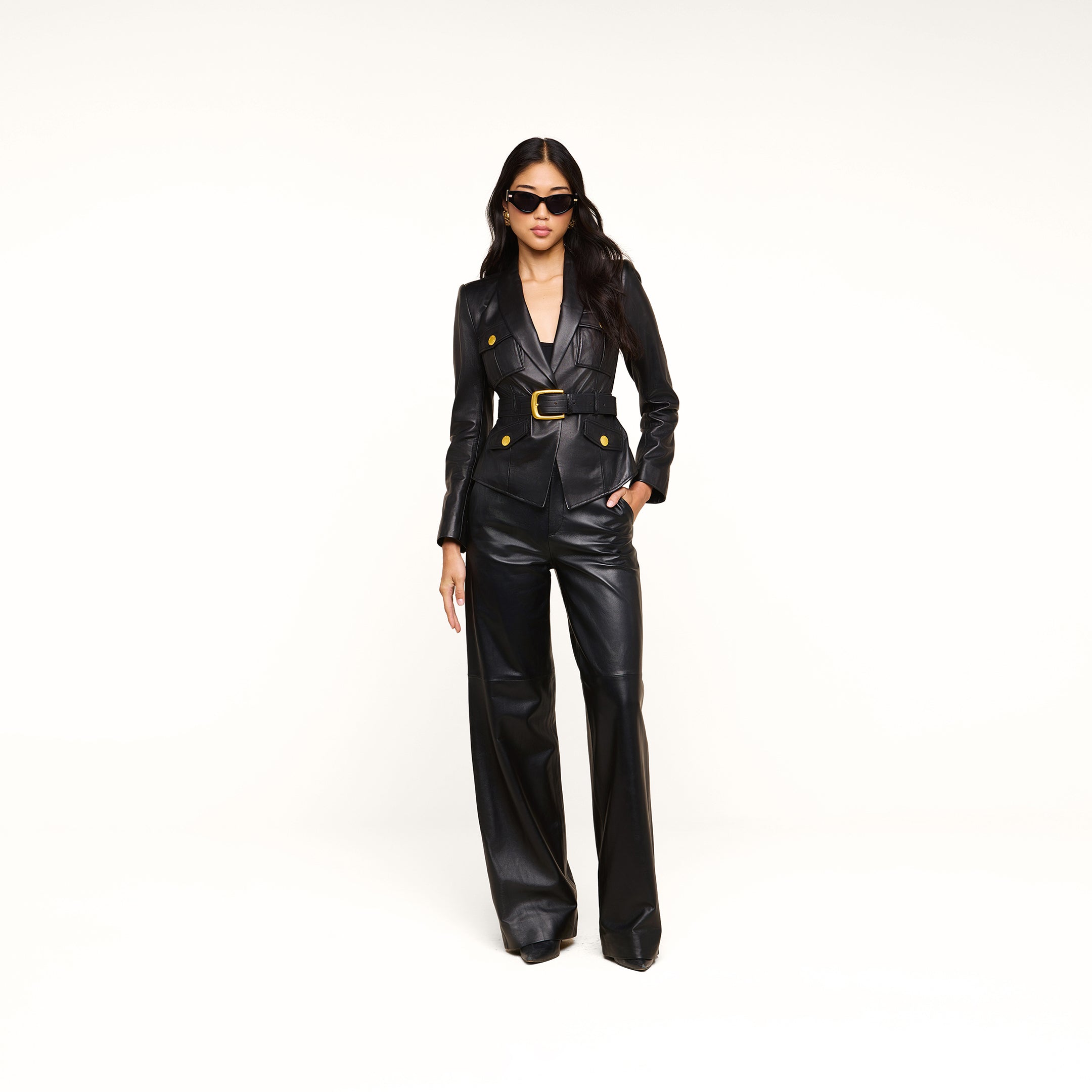 A woman wearing the Wisteria Leather Blazer, featuring a fitted black leather design with gold buttons and a wide belt, stands confidently against a plain white background, accessorized with sunglasses.