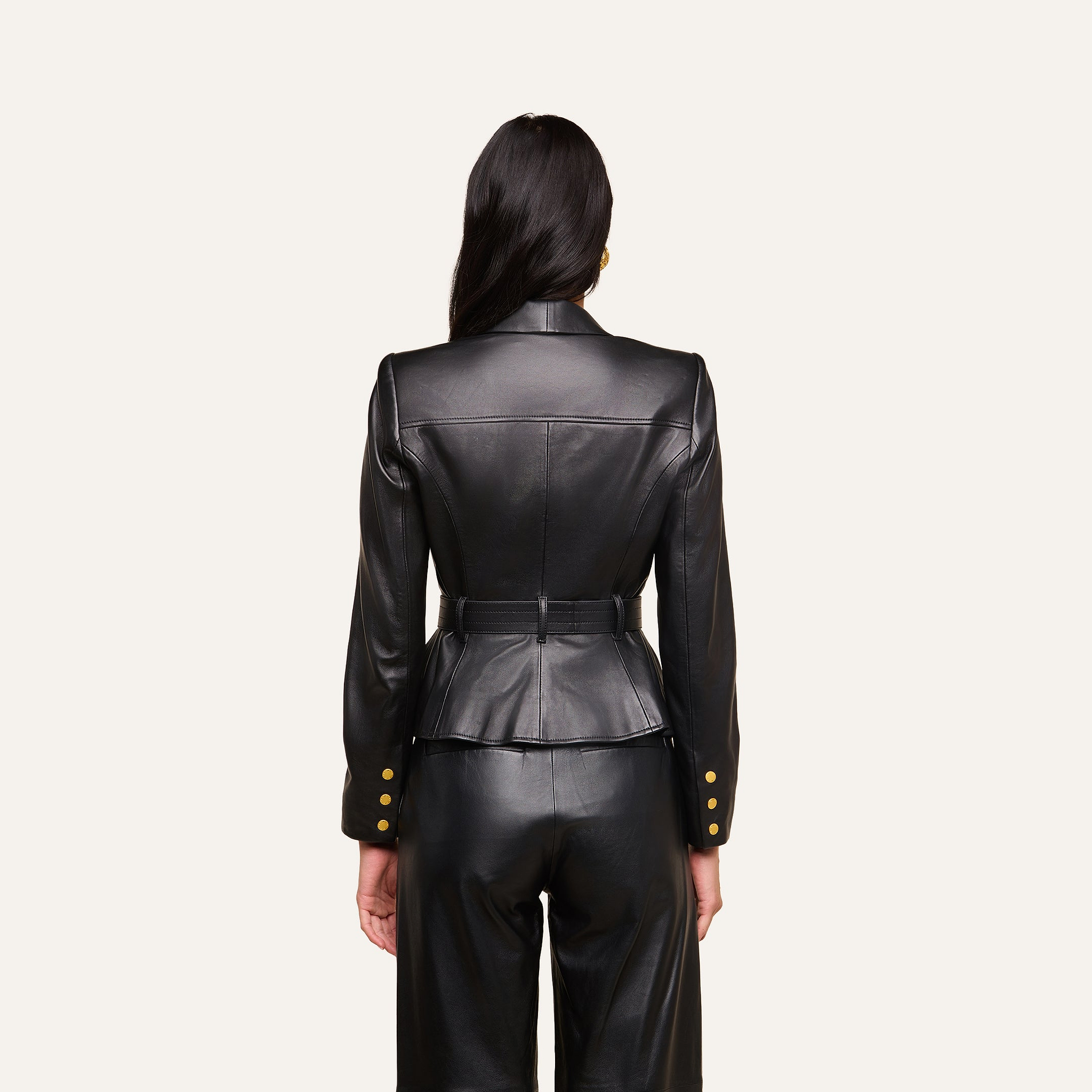 A woman with long dark hair, seen from behind, wears the fitted Wisteria Leather Blazer in black, paired with matching pants. The blazer features gold buttons on the cuffs and a belted waist. The background is plain and light-colored.