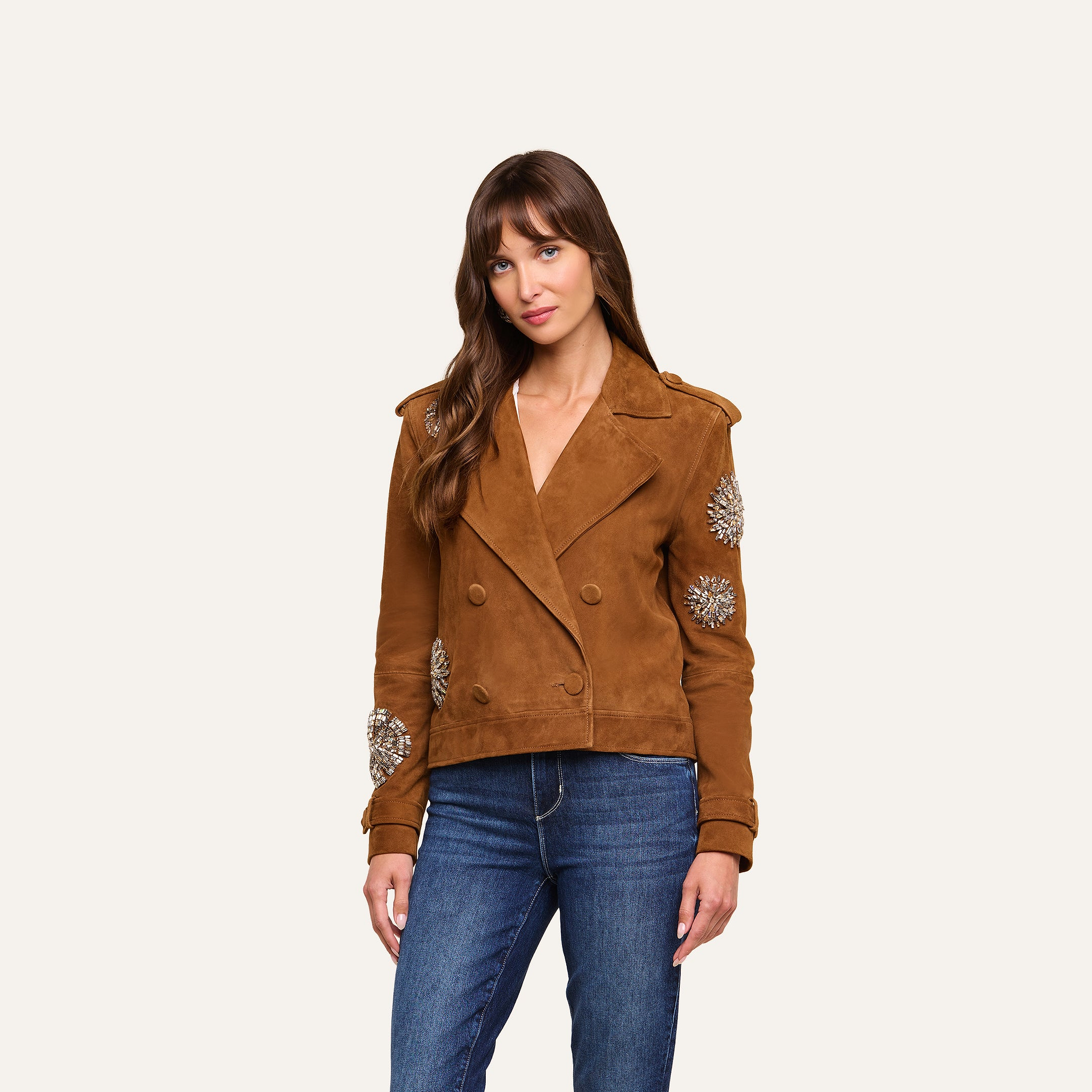 A woman wearing the Xoey Cropped Crystal Trench Coat— a brown suede double-breasted jacket adorned with crystal-embellished sleeves—paired with blue jeans, stands against a plain light background.