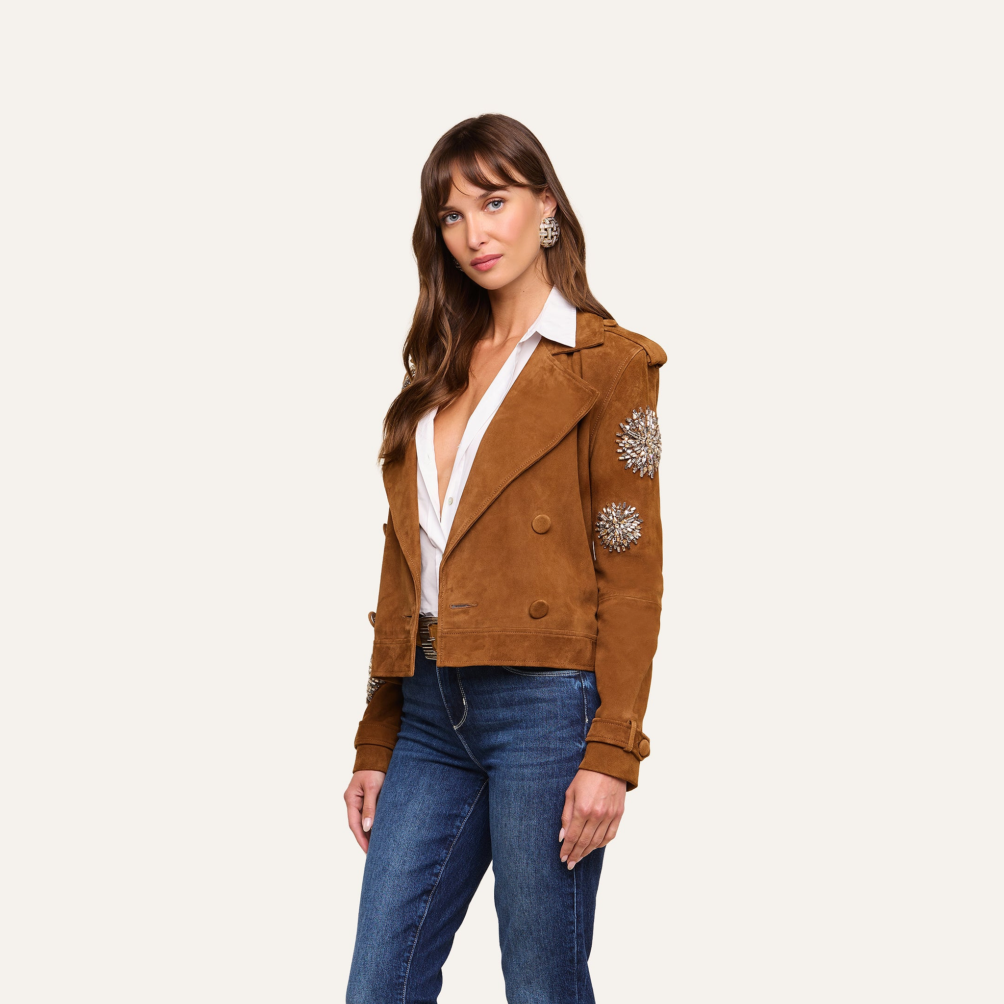 A woman models the Xoey Cropped Crystal Trench Coat—featuring crystal-embellished sleeves—over a white blouse and blue jeans, standing against a plain, light background for a chic, Fall 2026-inspired look.