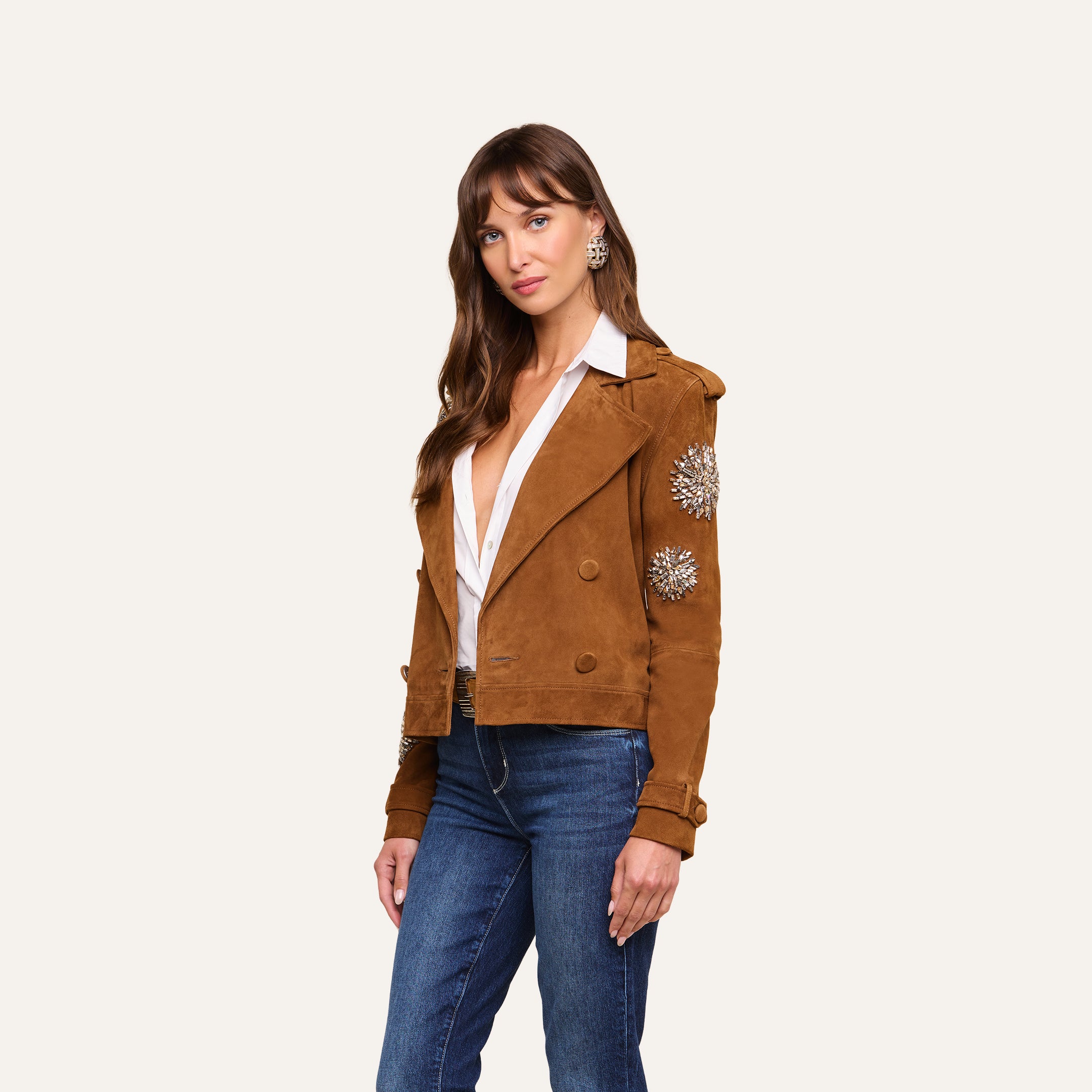 A woman models the Xoey Cropped Crystal Trench Coat—featuring crystal-embellished sleeves—over a white blouse and blue jeans, standing against a plain, light background for a chic, Fall 2026-inspired look.