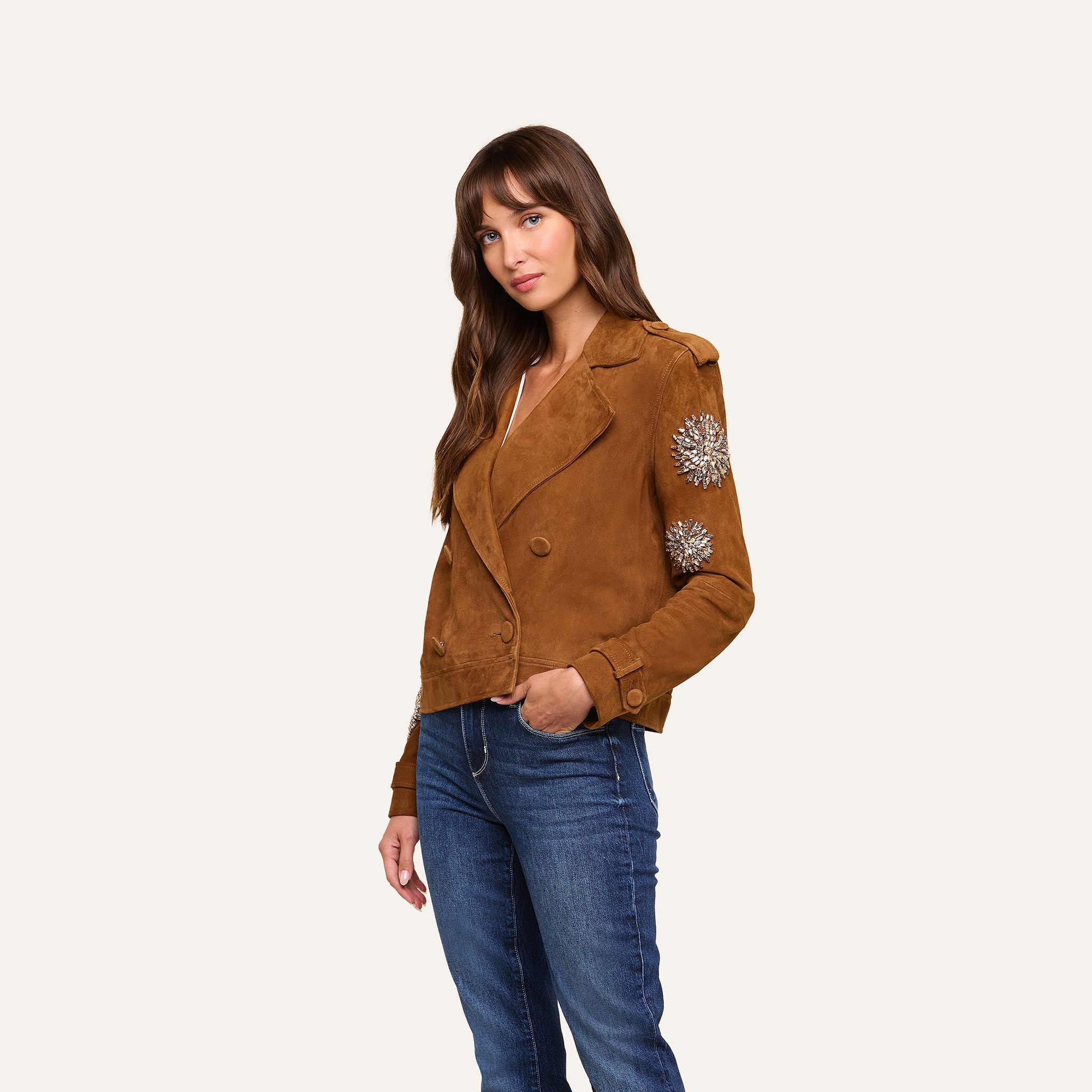 A woman stands against a plain light background, modeling the Xoey Cropped Crystal Trench Coat—its suede finish and crystal-embellished sleeves paired effortlessly with blue jeans—for a Fall 2026 Fashion Week-inspired look.