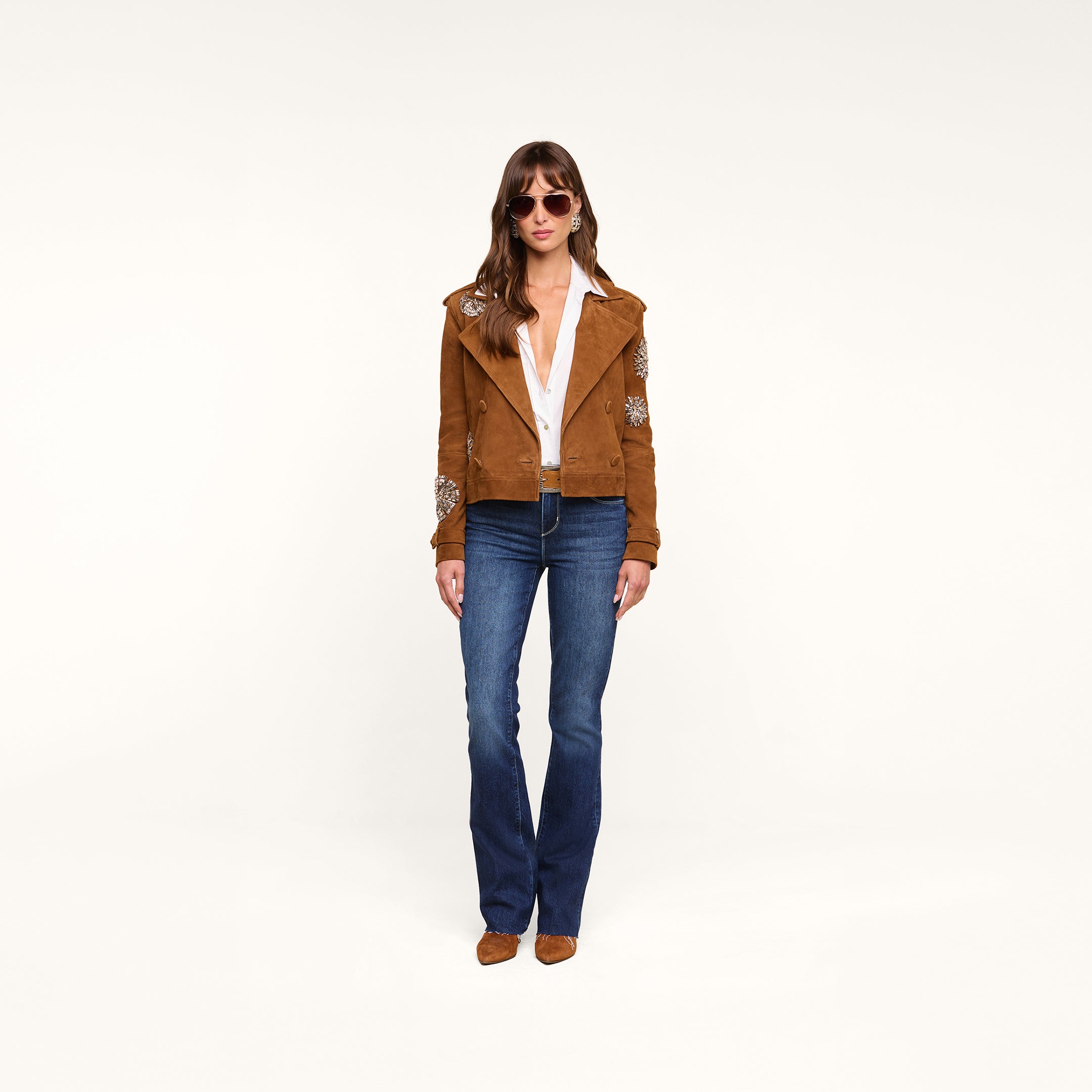 A woman wearing sunglasses, the Xoey Cropped Crystal Trench Coat, a white blouse, blue jeans, and brown shoes stands against a plain white background.