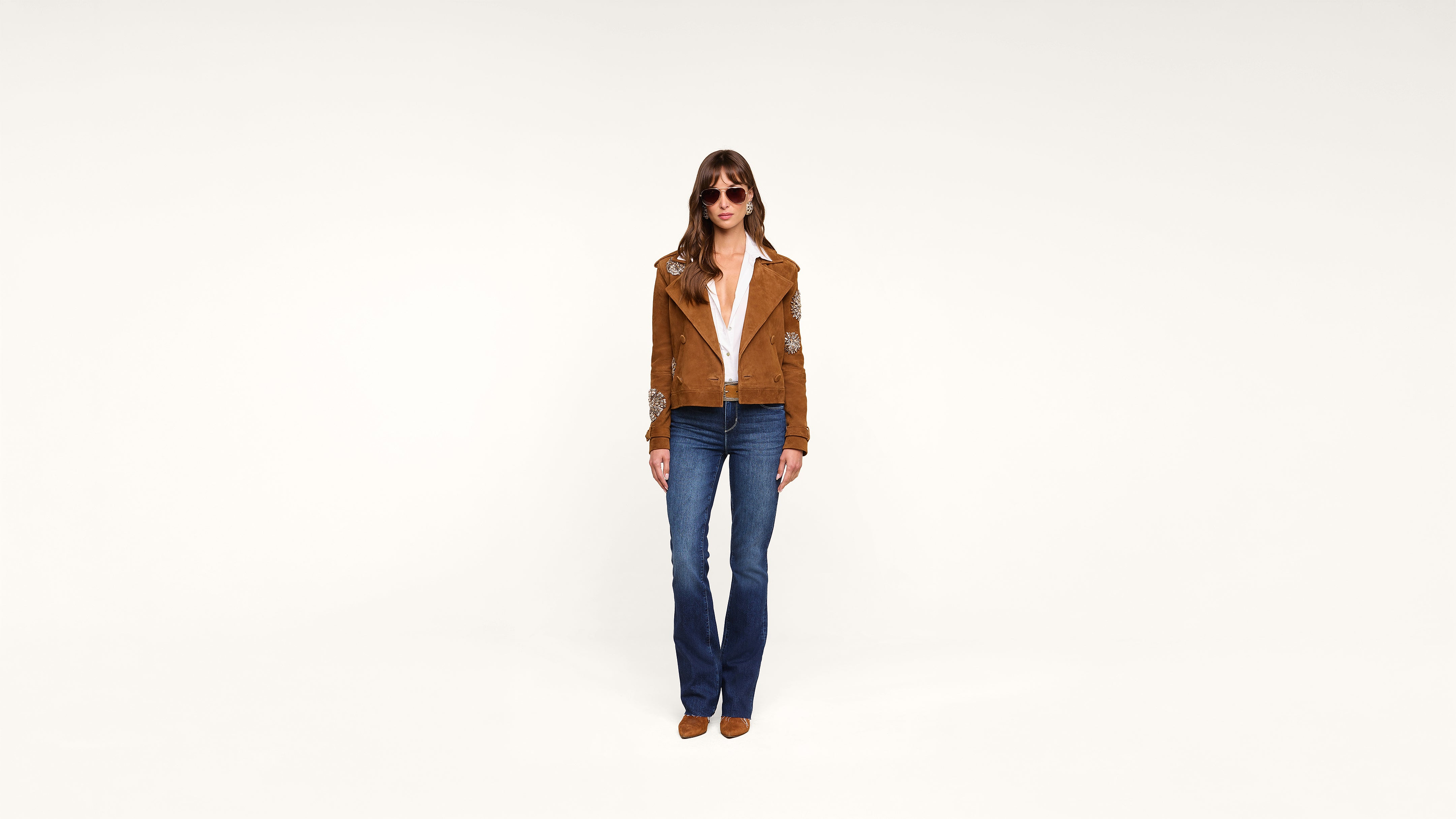 A person wearing sunglasses, a brown jacket over a white shirt, blue jeans, and brown shoes stands against a plain white background.