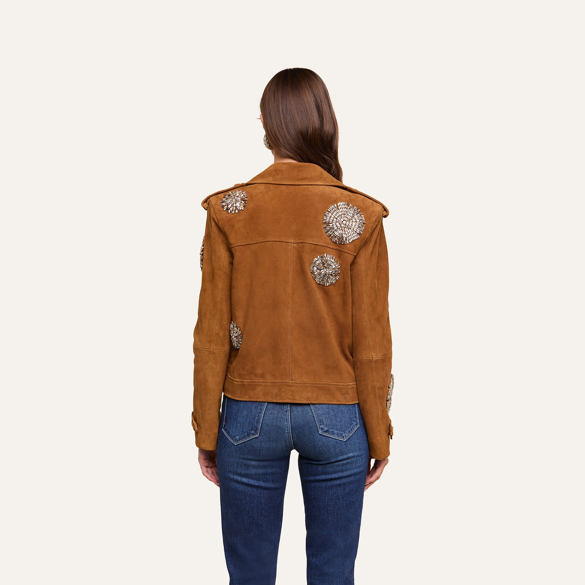 A person with long brown hair, wearing blue jeans and the Xoey Cropped Crystal Trench Coat—featuring decorative white floral patterns—is standing facing away from the camera against a plain light background, in a look inspired by Fall 2026 Fashion Week.