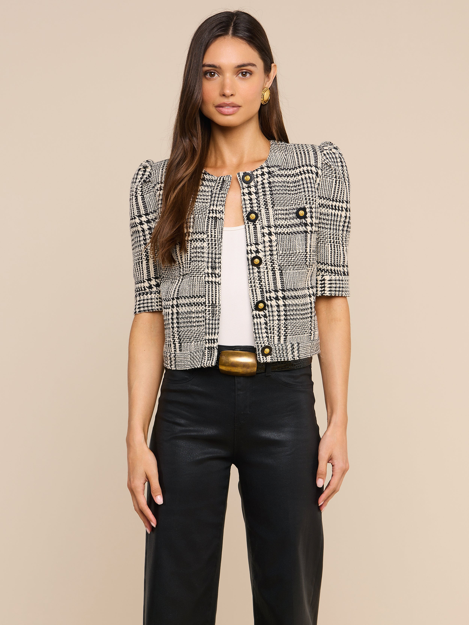 A woman stands against a plain background wearing the Allegra Cropped Tweed Jacket—a black-and-white plaid short-sleeve tweed piece with gold buttons—layered over a white top, paired with black high-waisted pants featuring a gold-buckled belt.