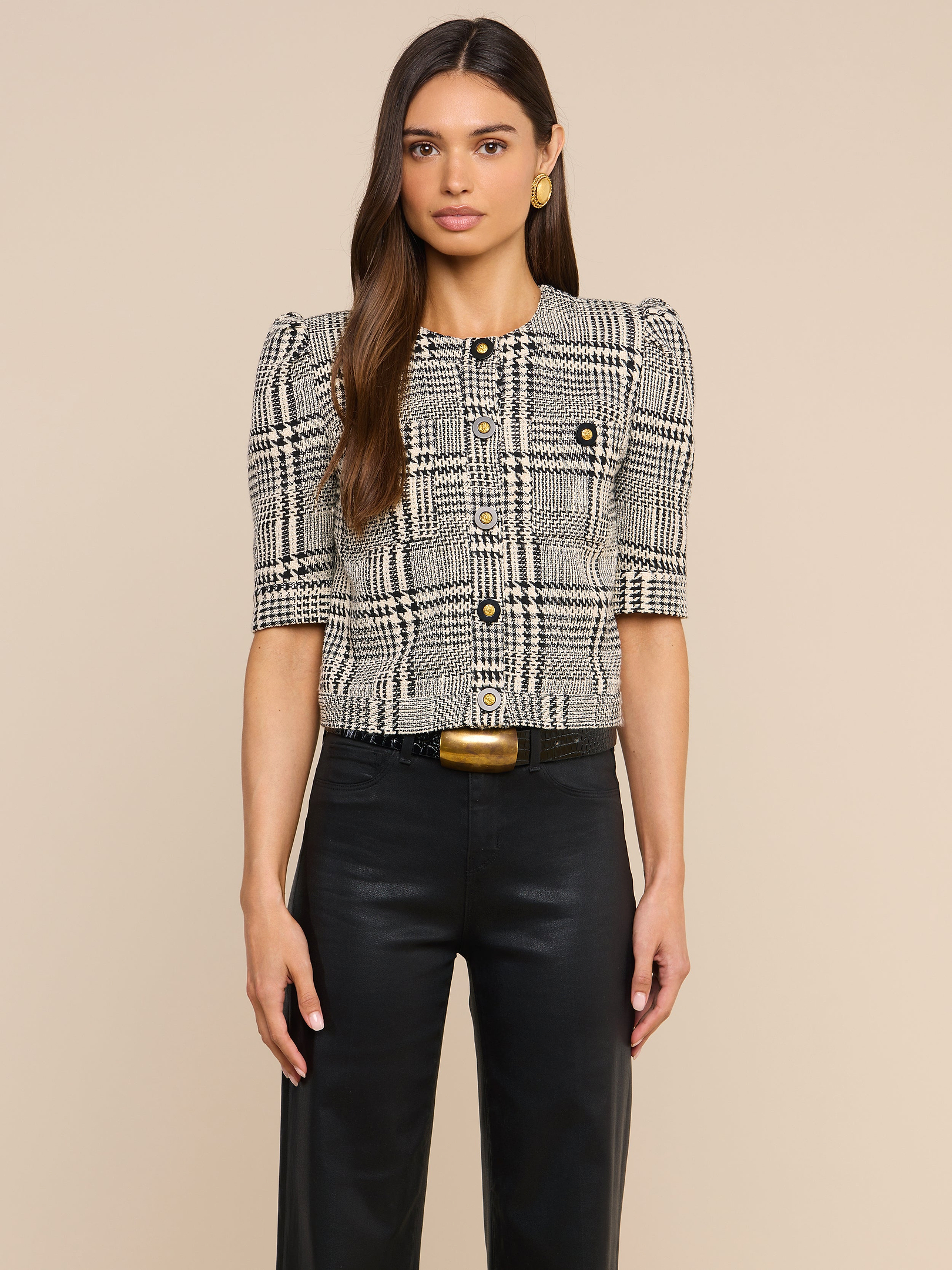 A woman with long brown hair is wearing the Allegra Cropped Tweed Jacket, which features a plaid pattern and short sleeves, paired with black pants, gold earrings, and a gold-buckled belt, standing against a plain light background.