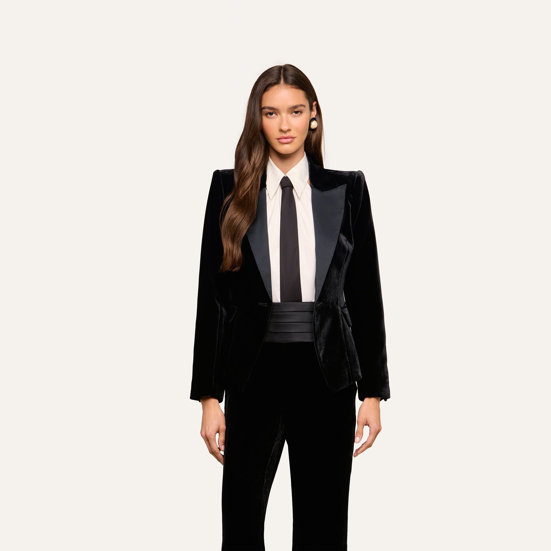 A woman wearing the Philippa Velvet Blazer with satin lapels, styled with a white dress shirt, black tie, and black cummerbund, stands against a plain light background. She has long, wavy hair and gazes forward.