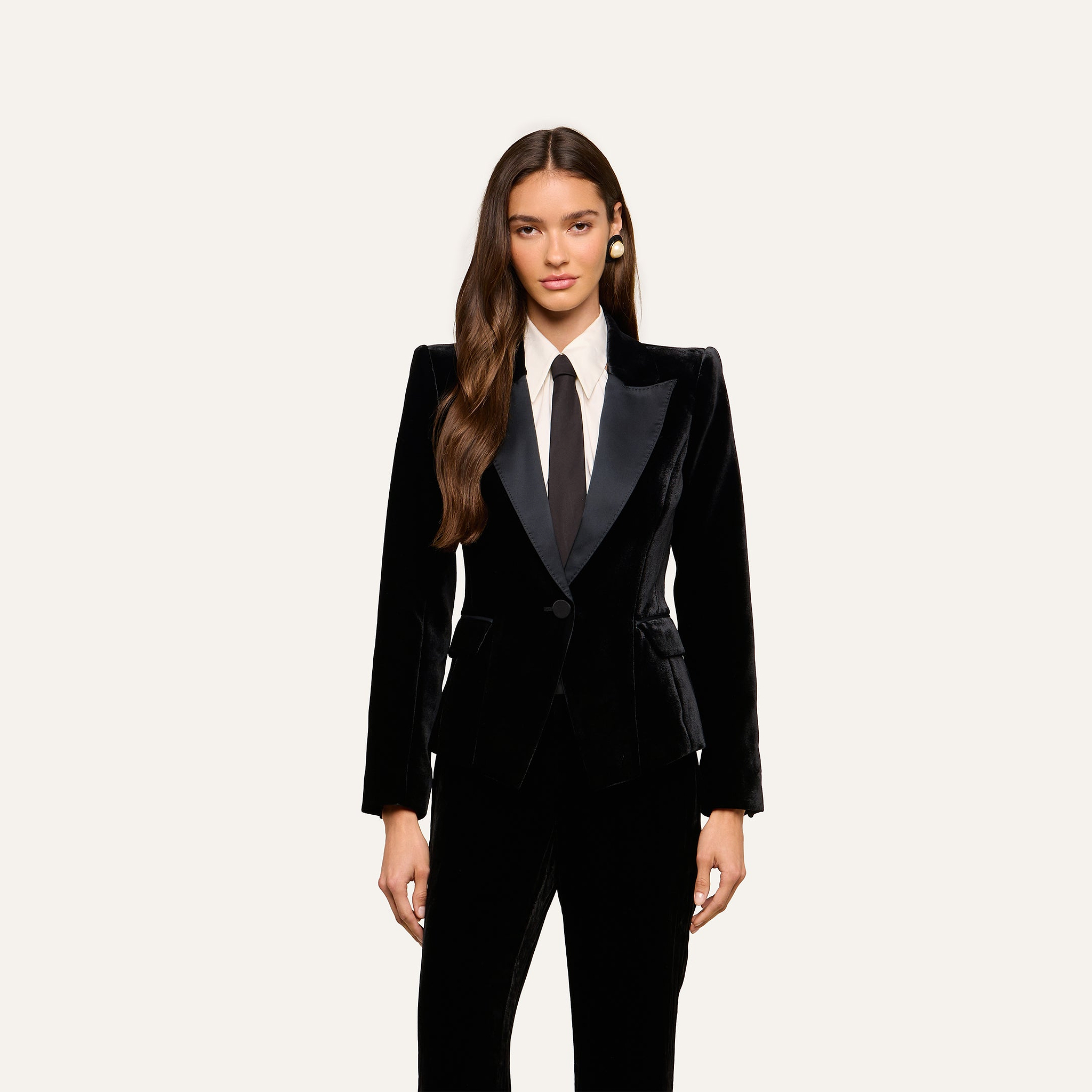 A woman with long brown hair wears the Philippa Velvet Blazer with satin lapels, paired with a white dress shirt and a black tie, standing against a plain light background.