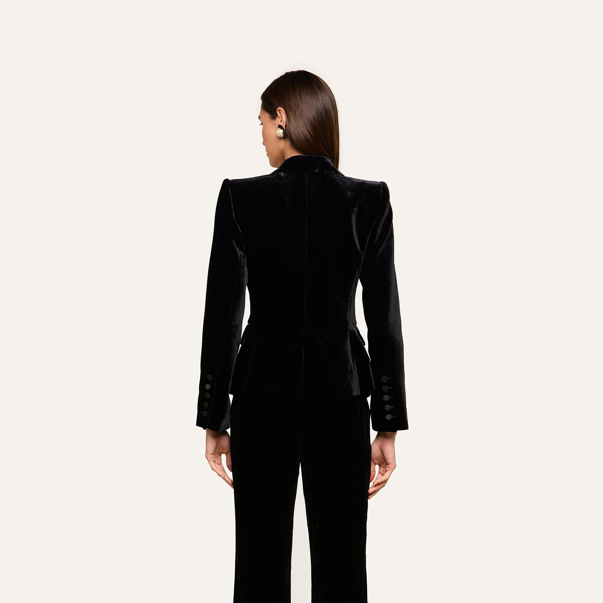 A woman stands with her back to the camera, wearing the fitted Philippa Velvet Blazer with satin lapels and matching black pants against a plain light background.