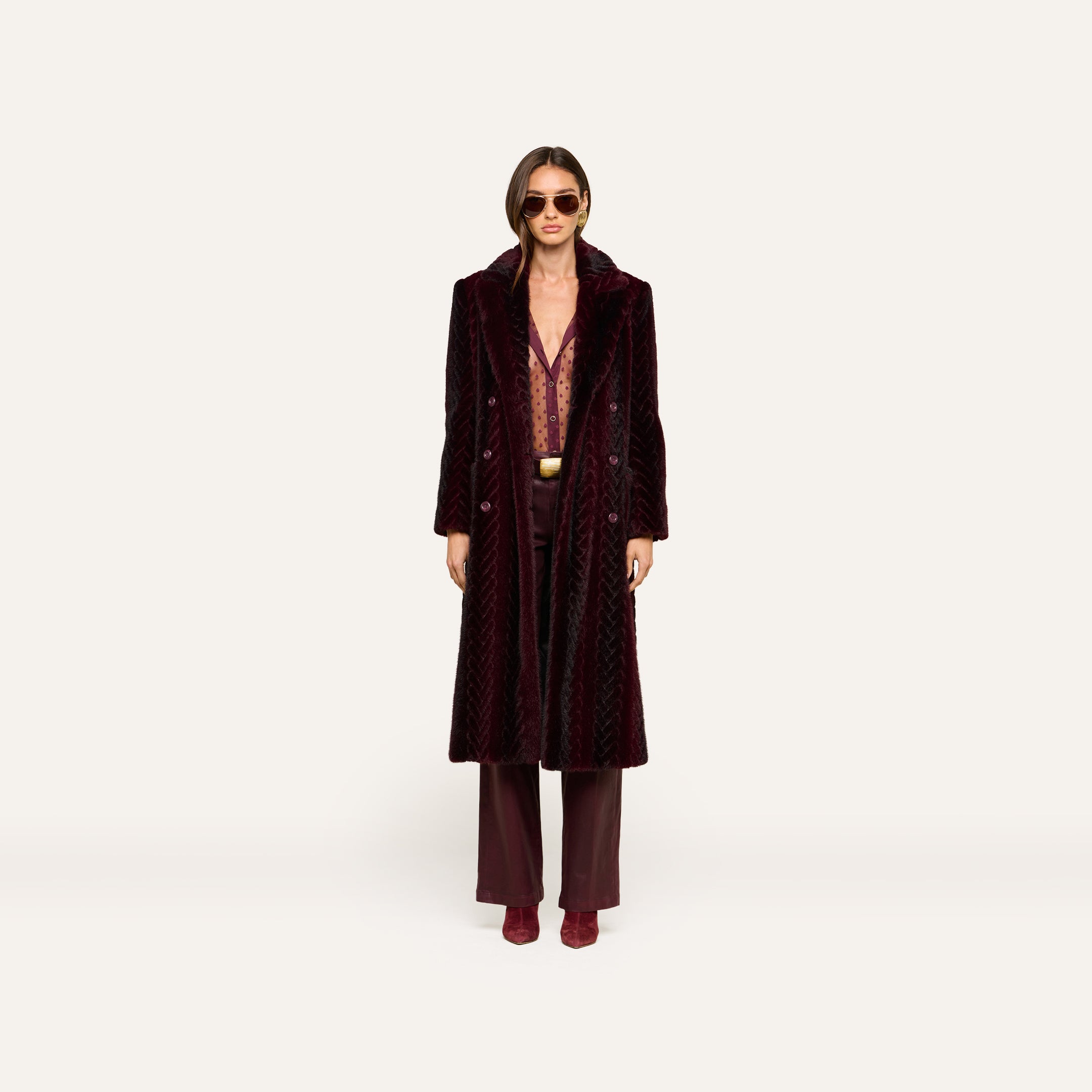 A person wearing the Icelyn Faux Fur Coat—a long burgundy velvet coat with faux fur accents—along with matching trousers, a patterned shirt, burgundy boots, and sunglasses stands against a plain light background.
