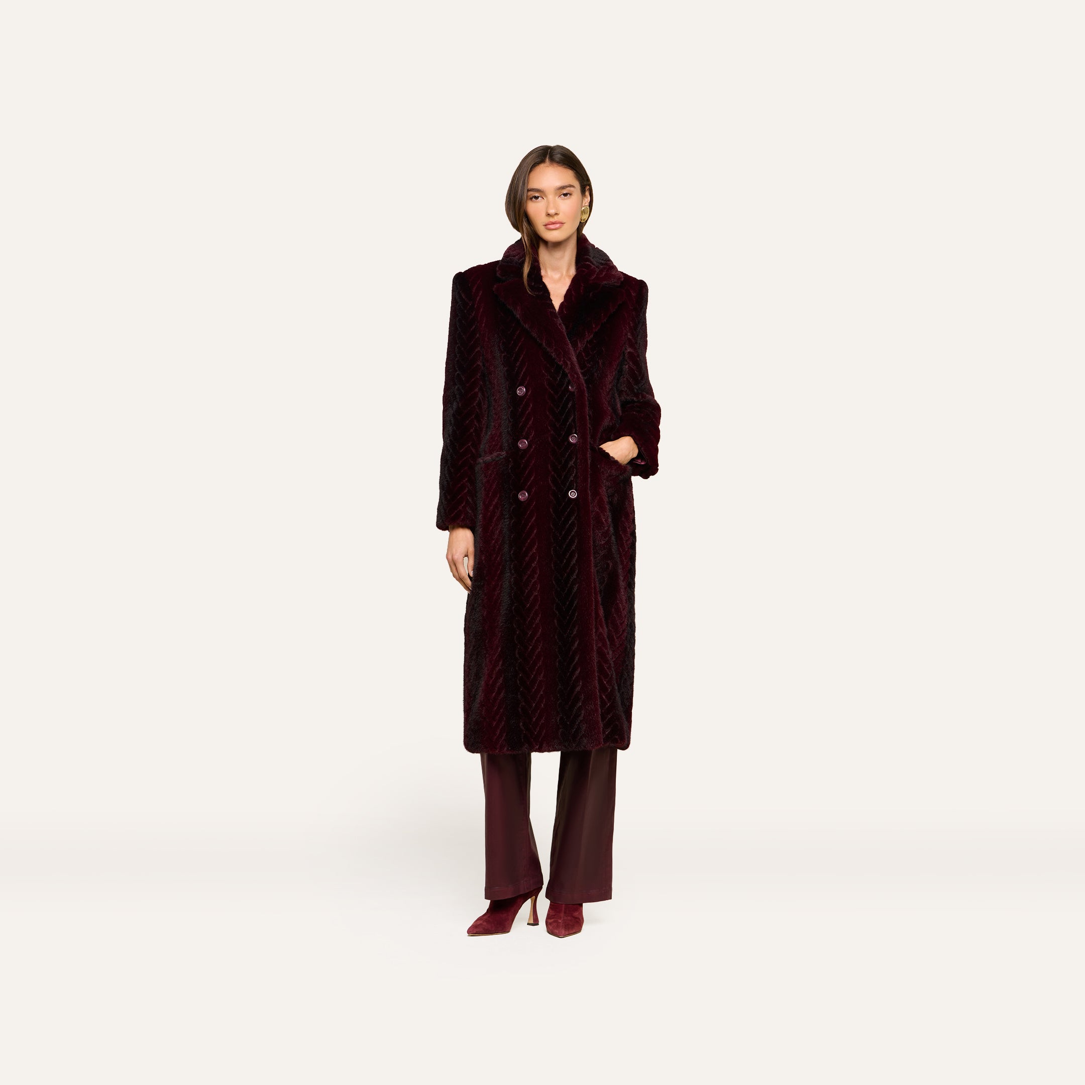 A woman wearing the Icelyn Faux Fur Coat—a long, dark burgundy velvet coat with a chevron pattern and faux fur accents—paired with matching pants and burgundy heels, standing against a plain white background.