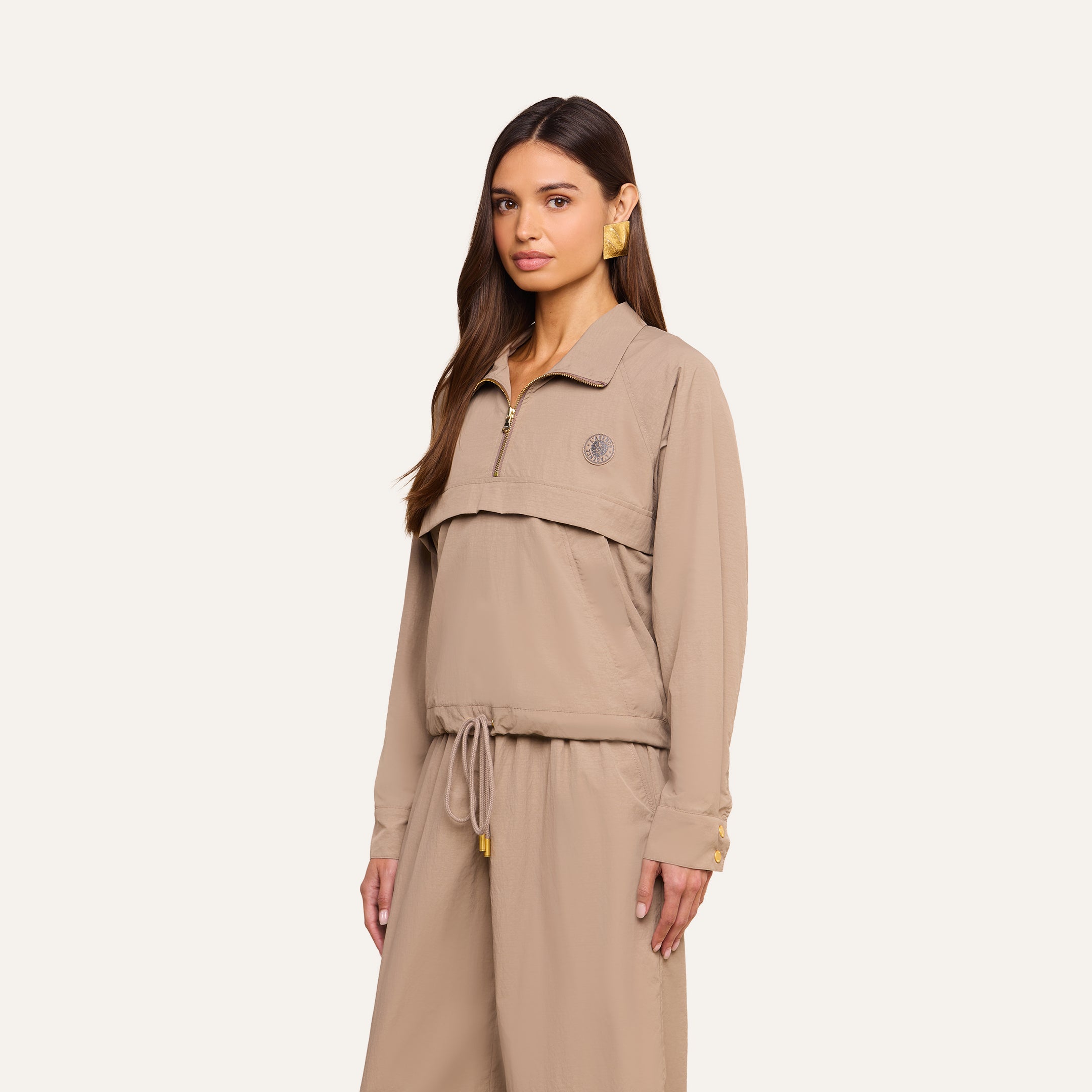 A woman with long brown hair wears the Vex Cropped Windbreaker in beige, paired with matching loose pants, standing against a plain light background and facing forward with a neutral expression.
