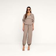A woman stands against a plain background wearing sunglasses, the Vex Cropped Windbreaker in light brown, matching jogger pants, and heeled sandals.