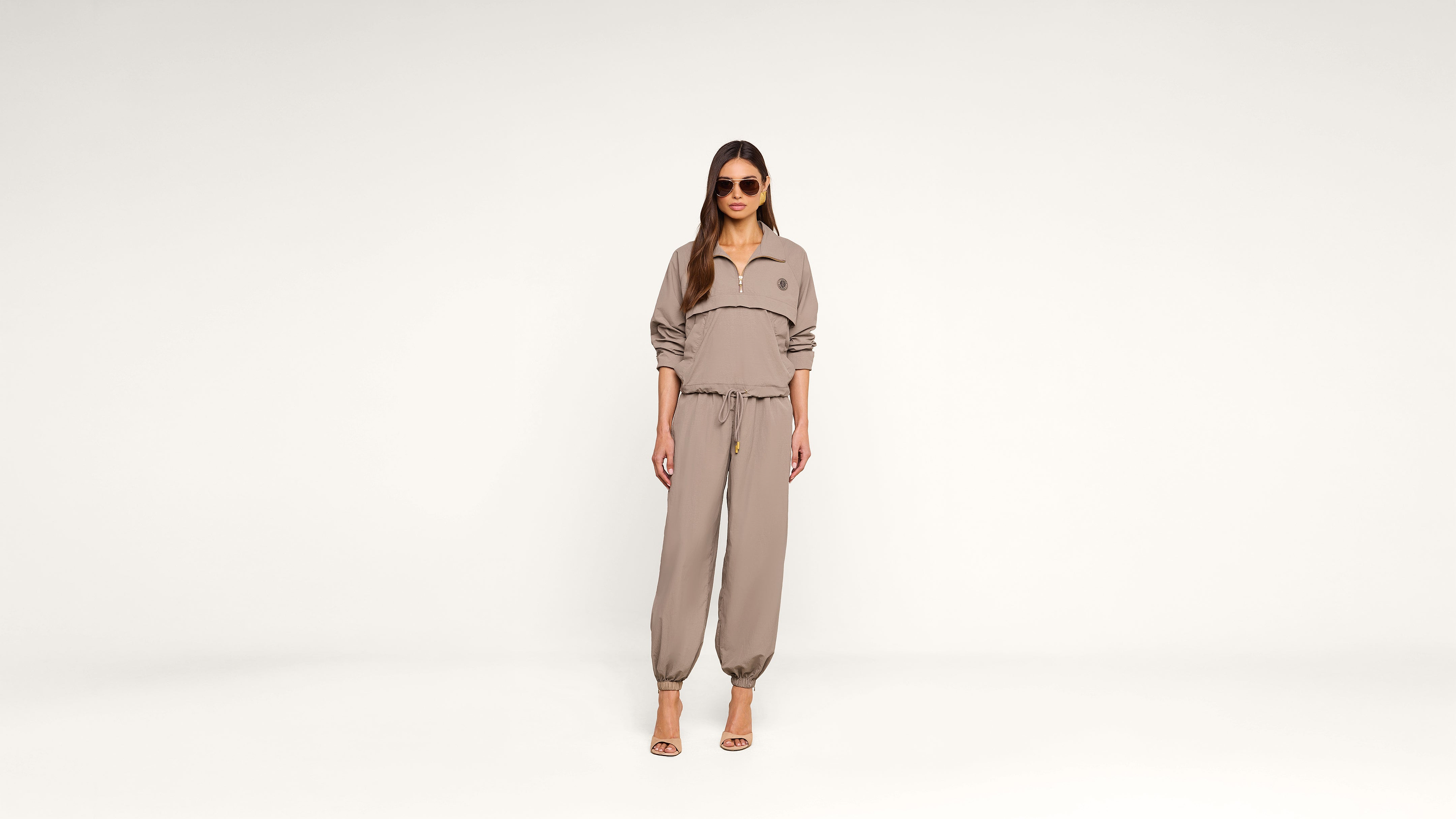 A woman wearing sunglasses, a taupe zip-up top, and matching jogger pants stands against a plain white background, looking straight ahead.