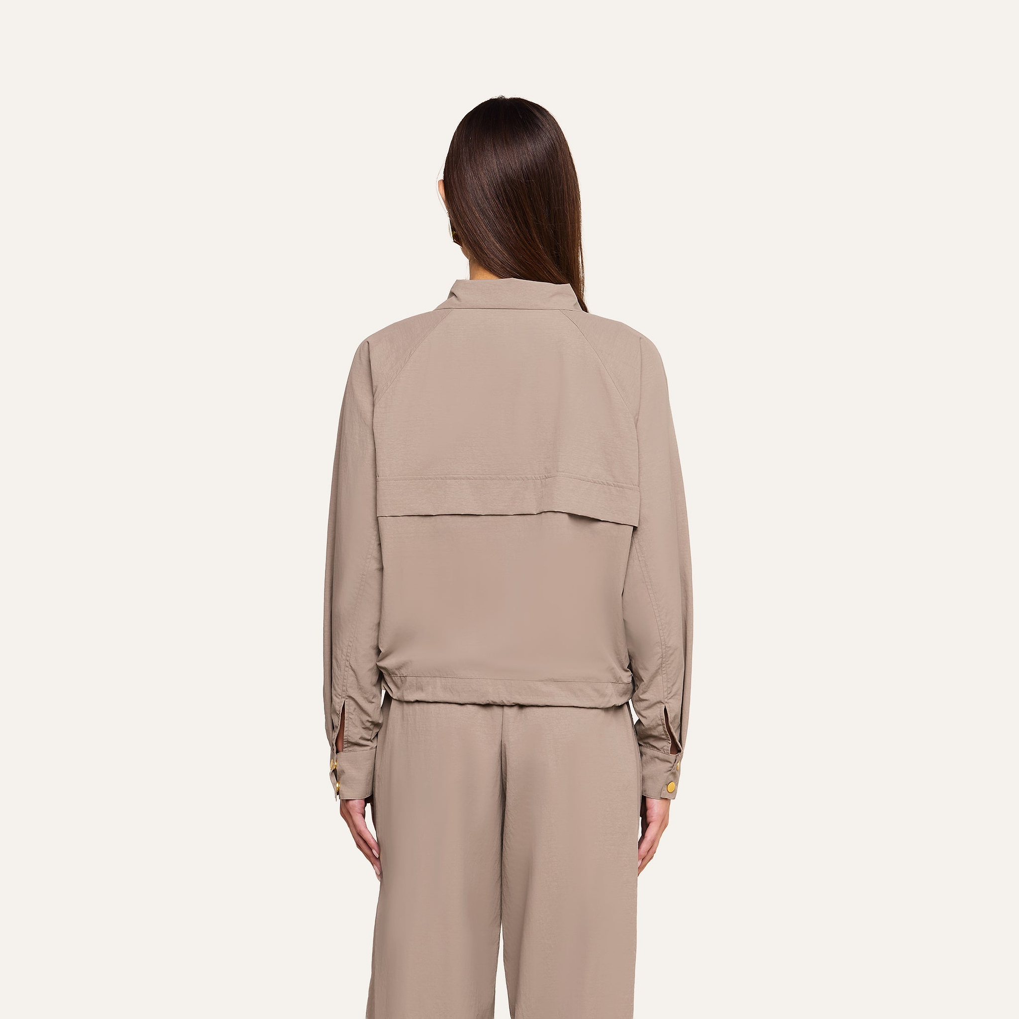 A person with straight brown hair, wearing the Vex Cropped Windbreaker in loose beige, along with matching pants, stands facing away from the camera against a plain light background.