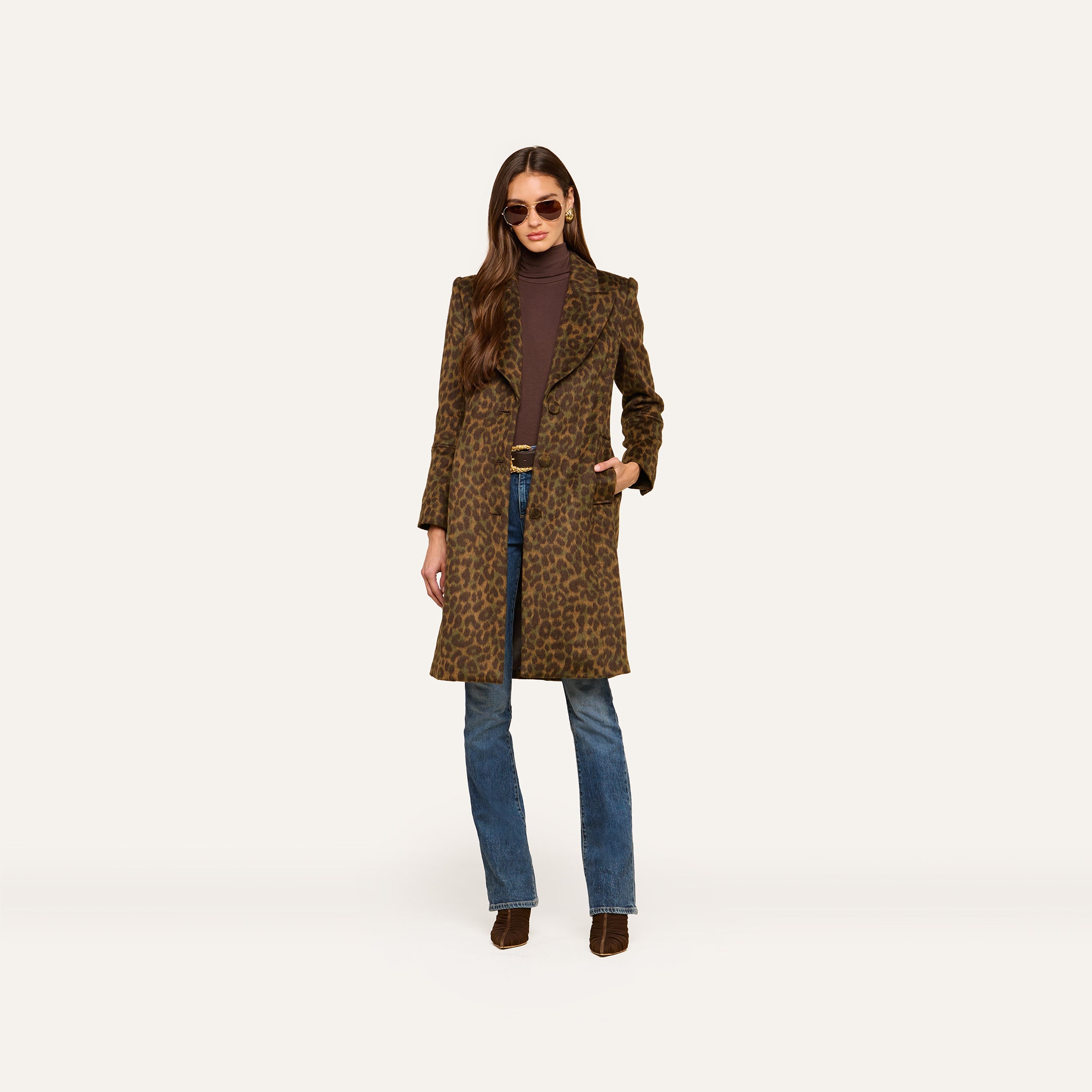 A woman stands against a plain light background wearing the Laney Coat, a long collared leopard print piece, paired with a brown turtleneck, blue jeans, and brown boots. She has long brown hair and sunglasses.