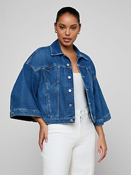 A woman wearing the Silas Cape Jacket, a cropped all-cotton denim jean shirt jacket, over a white top and white pants stands against a plain light background.