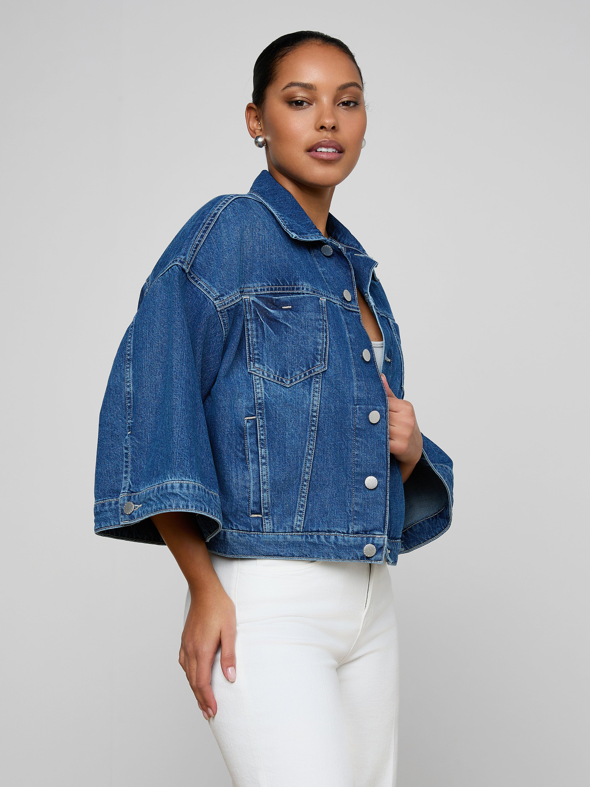 A woman poses in the Silas Cape Jacket—an all-cotton denim shirt jacket with cape sleeves—paired with white pants against a plain, light background.