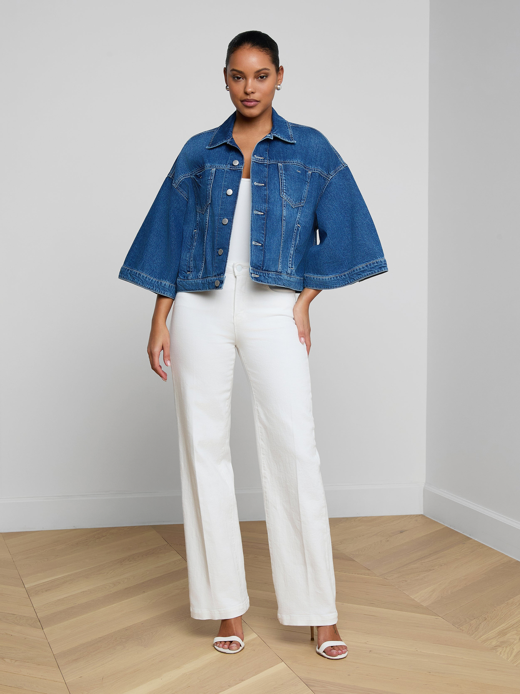 A woman stands against a plain, light background wearing the Silas Cape Jacket—an all-cotton denim shirt jacket featuring cape sleeves—layered over a white top and wide-leg white pants with heeled sandals.