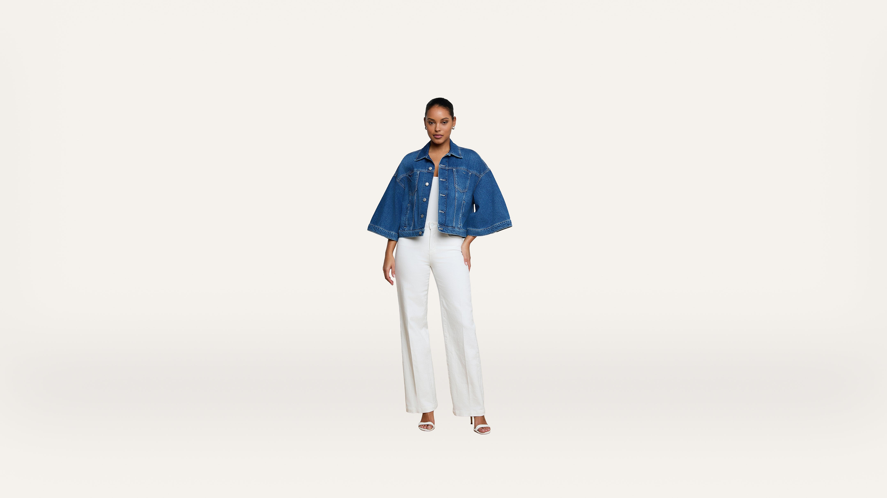 Woman stands facing forward, wearing a blue denim jacket over a white top and white wide-leg pants, with white heeled sandals, on a plain white background.