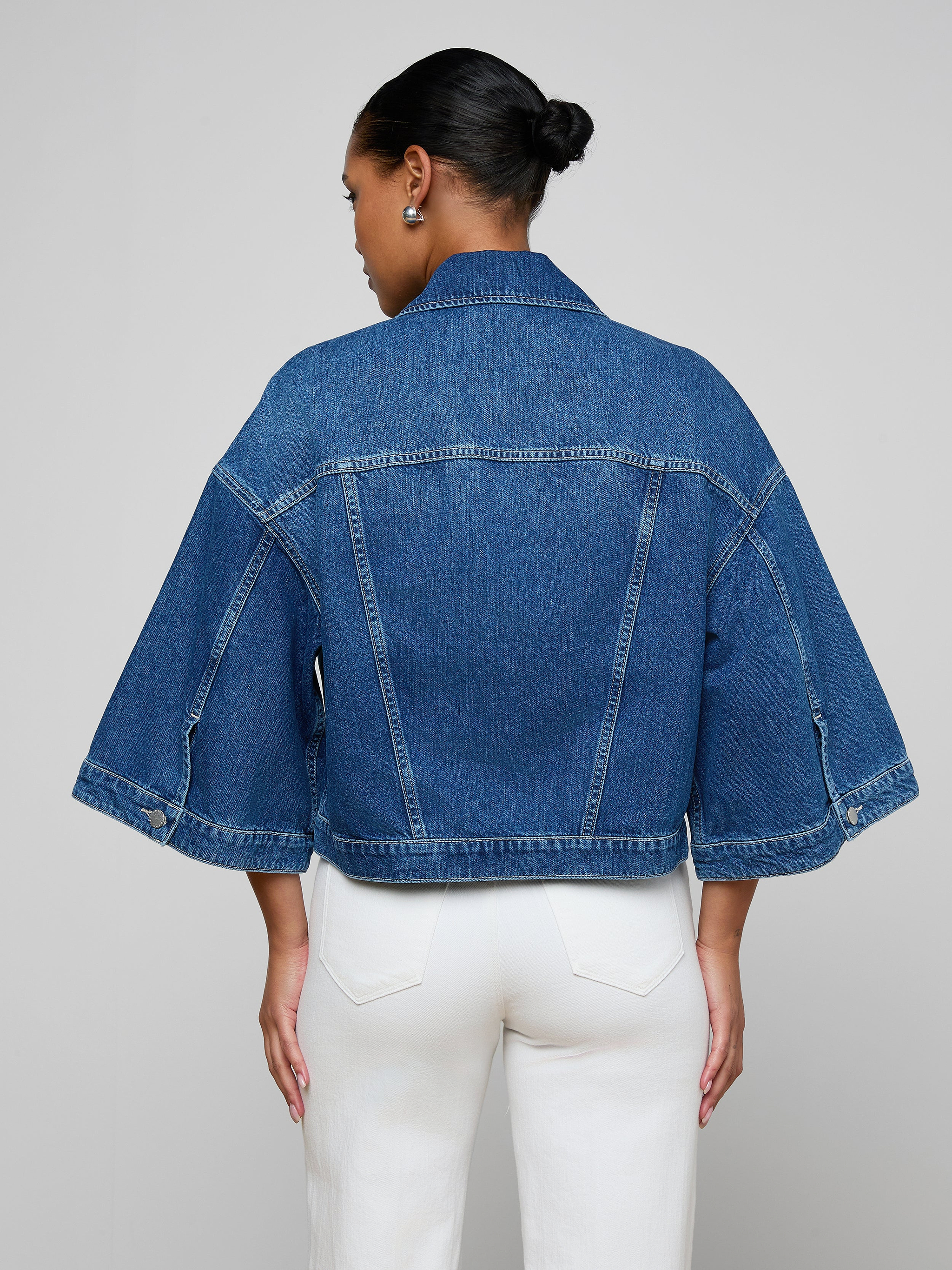 Wearing the Silas Cape Jacket—a 100 cotton denim shirt jacket with cape sleeves—paired with white pants, a person with their hair in a bun stands facing away from the camera against a simple light background.