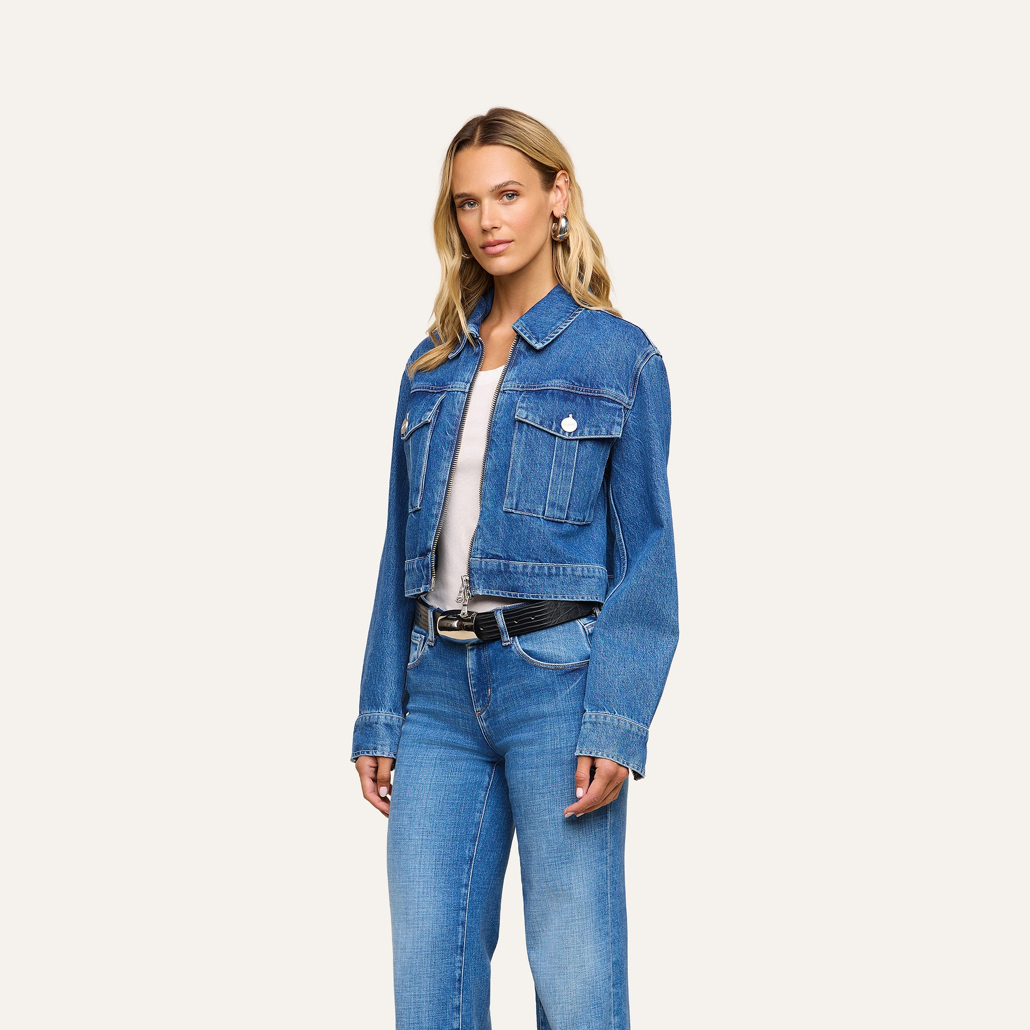 A woman with long blonde hair stands facing forward against a plain, light-colored background, wearing the Micah Oversized Jean Jacket and jeans. The jacket features an oversized fit in blue denim, and she is dressed casually with one arm relaxed by her side.