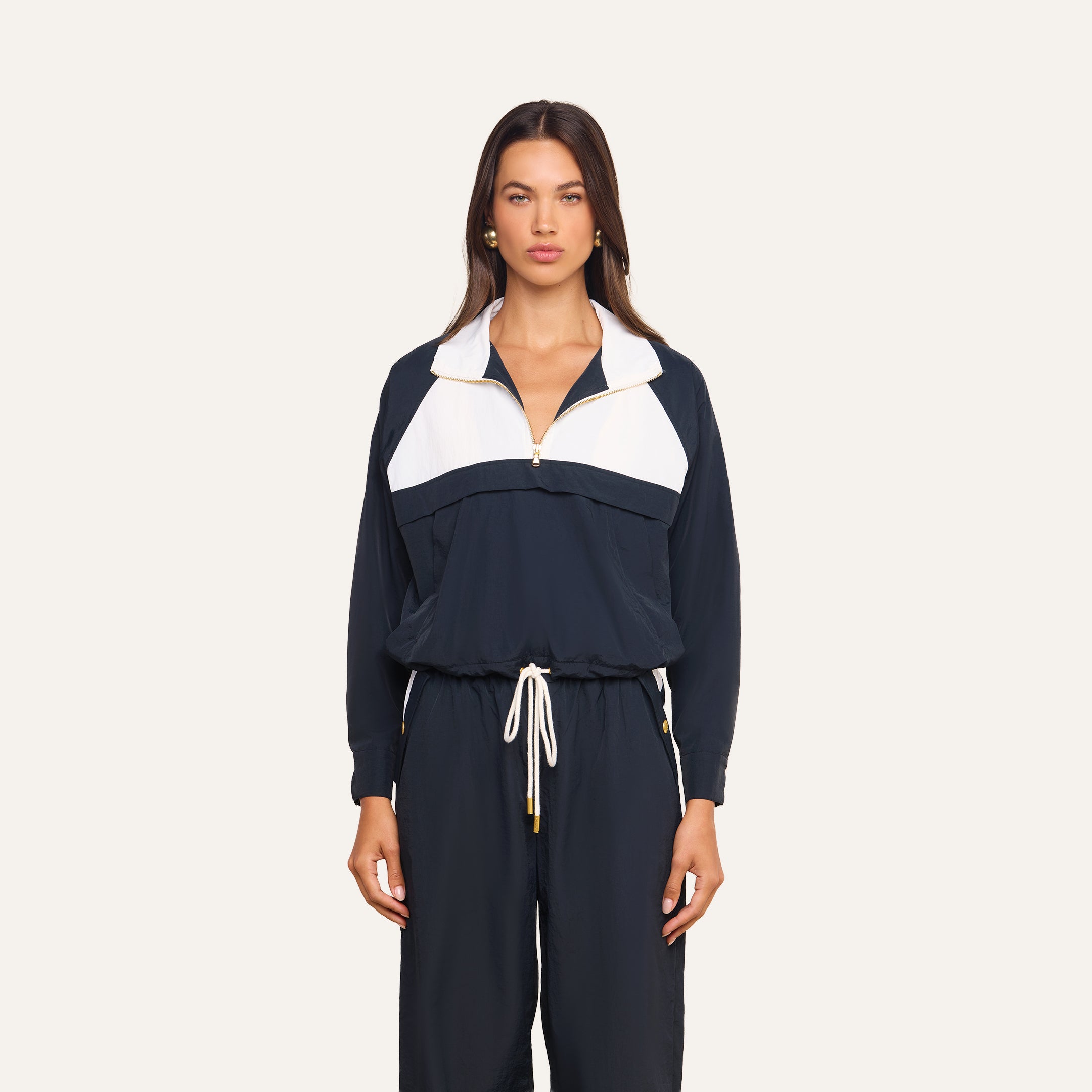 A woman stands facing forward, wearing the Vex Cropped Windbreaker in navy blue and white with matching drawstring pants, against a plain light background.