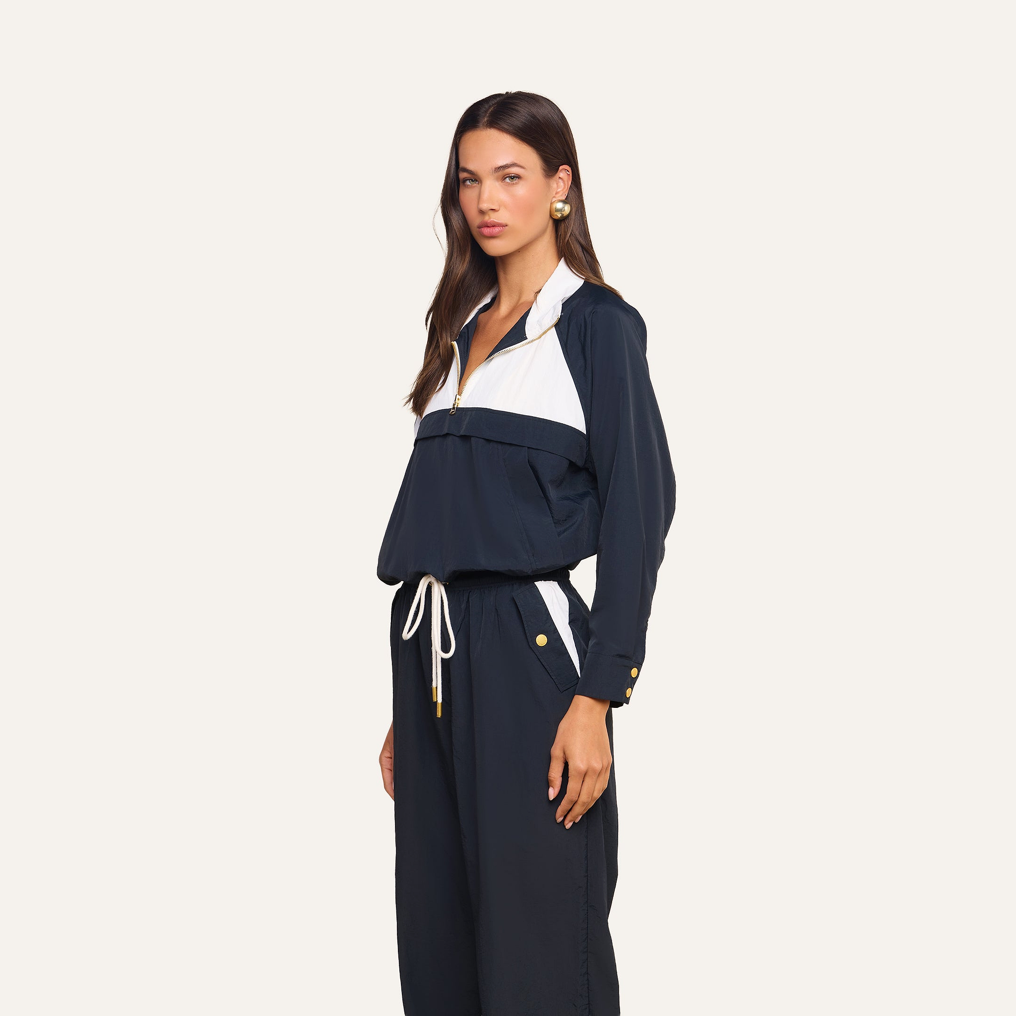 A woman wears the Vex Cropped Windbreaker, a navy blue and white long-sleeve activewear jacket, paired with matching drawstring pants while standing against a plain background.