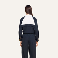 A woman with straight brown hair, wearing a black jumpsuit featuring a white panel on the back, stands facing away against a plain light background, paired with the Vex Cropped Windbreaker.