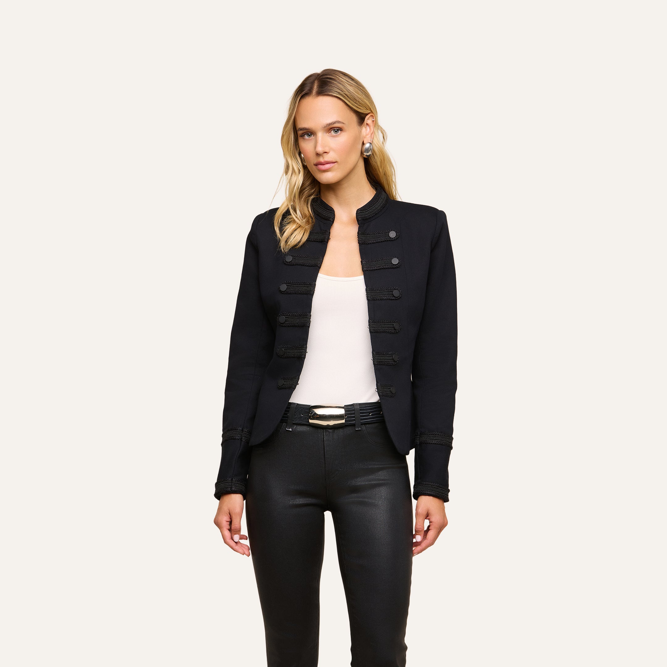 A woman wearing the Victory Jacket, a vintage-inspired black military-style piece, layered over a white top and paired with black pants and a belt, stands against a plain light background.