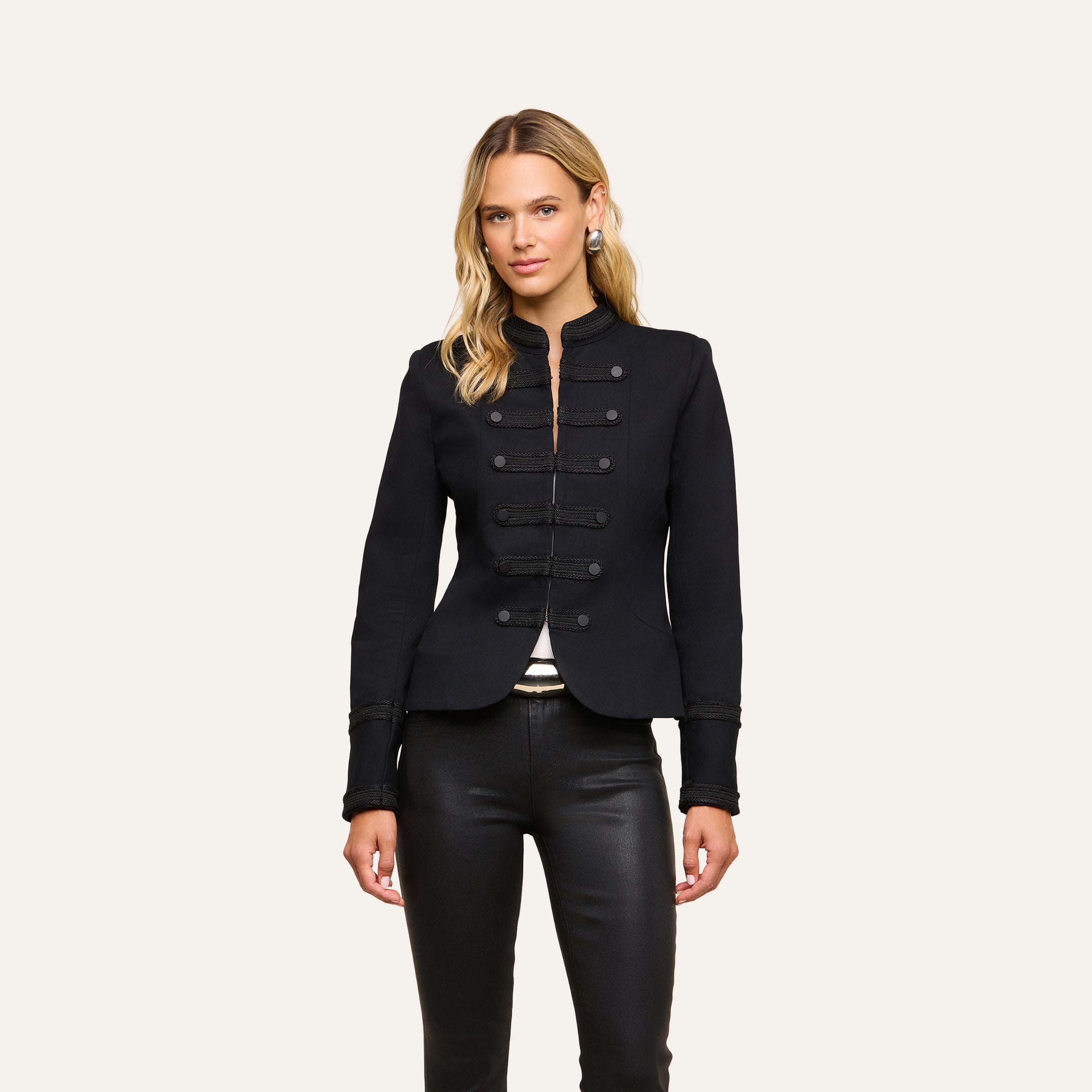 A woman is wearing the Victory Jacket, a fitted black Victorian-style piece with decorative buttons, paired with black leather pants and standing against a plain, light background.